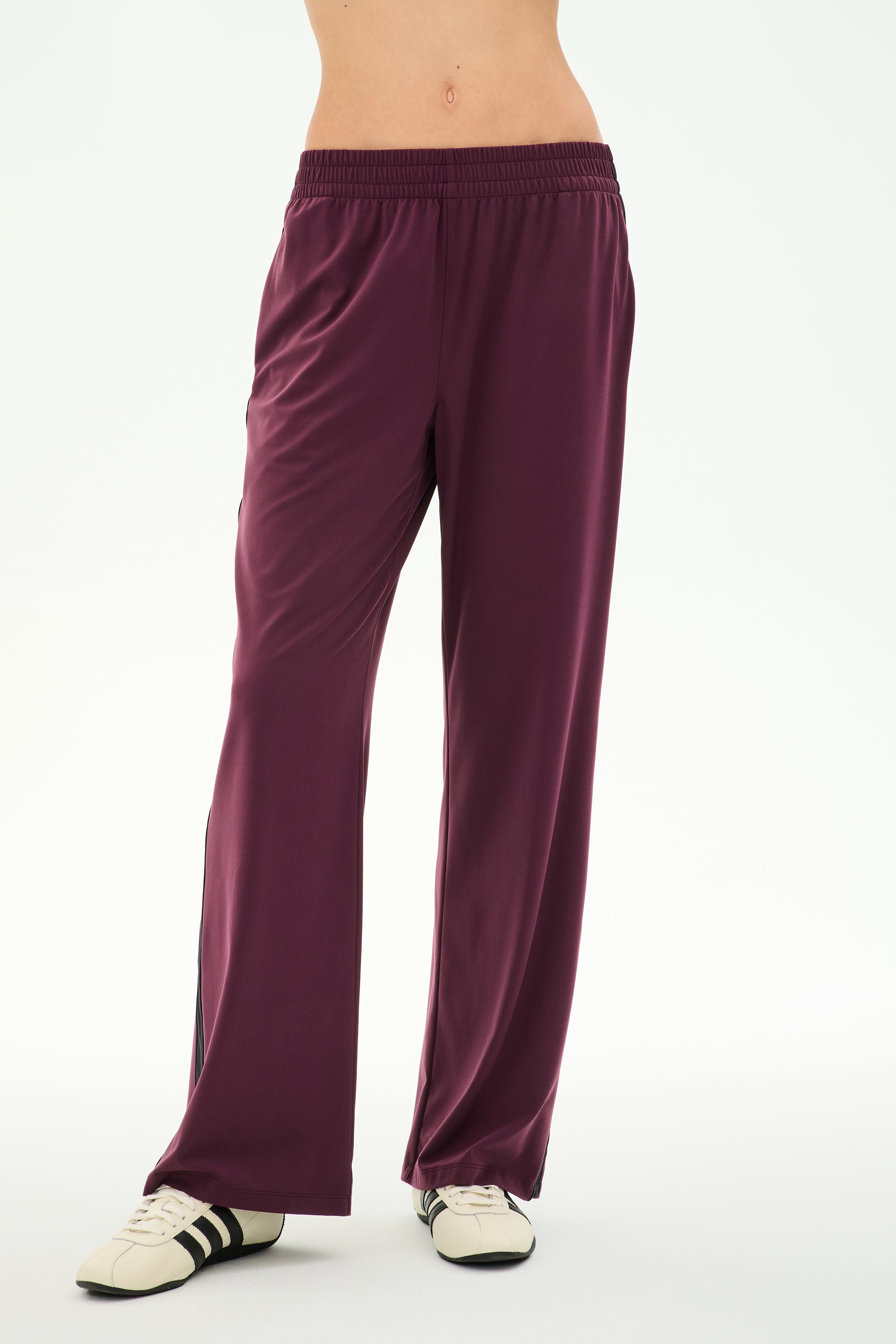 A person wearing the SPLITS59 Maxie Rigor Track Pant in Dark Cherry/Black, crafted from a soft nylon-spandex blend, paired with white sneakers featuring black stripes, stands against a plain white background.