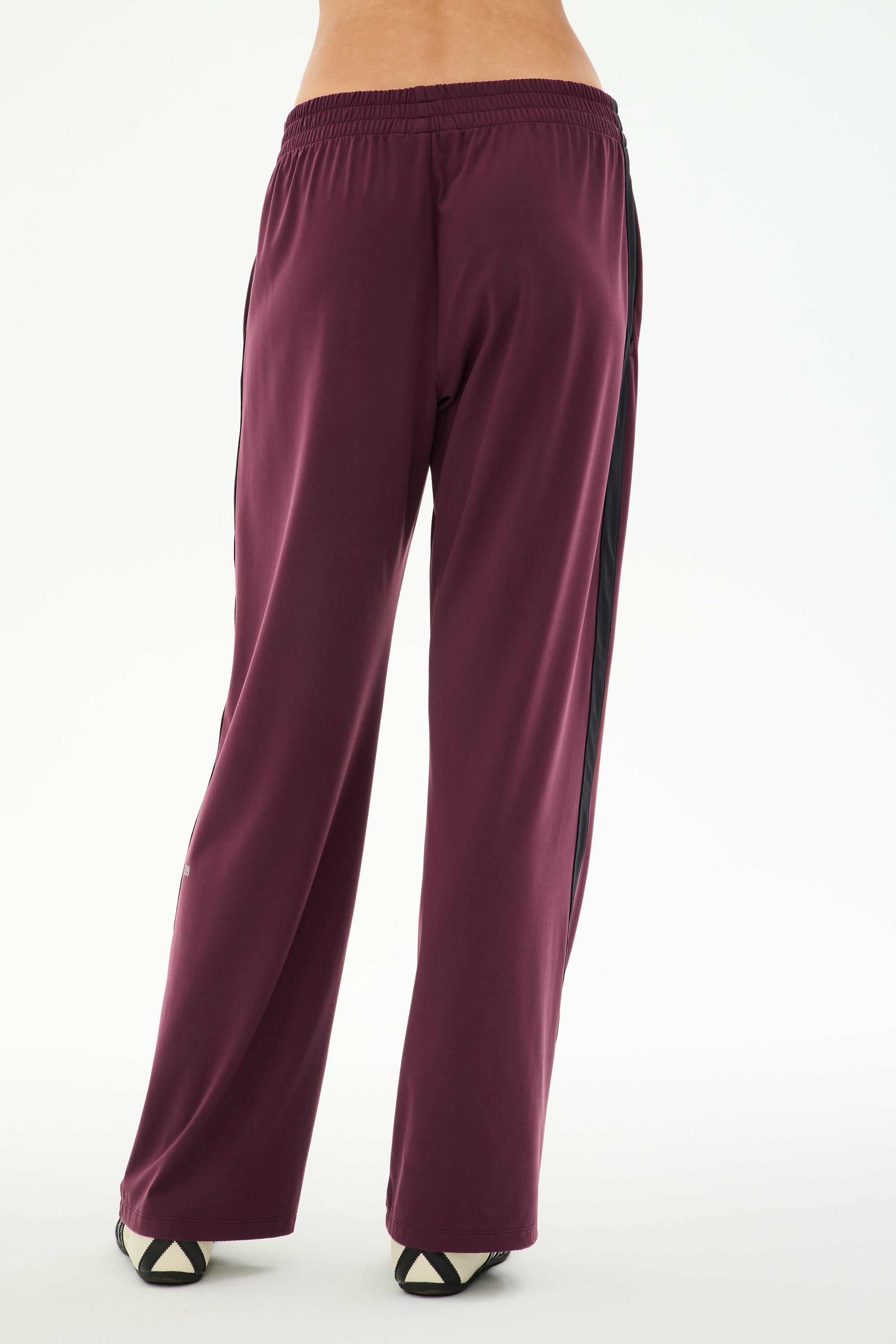 A person is seen from the back against a plain white background wearing SPLITS59’s Maxie Rigor Track Pant in Dark Cherry/Black, featuring a loose fit and black side stripes.