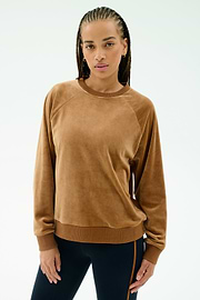 A woman with braided hair wears the SPLITS59 Joy Velour Crewneck in Honey, paired with dark side-striped stretch pants, standing against a plain light background.