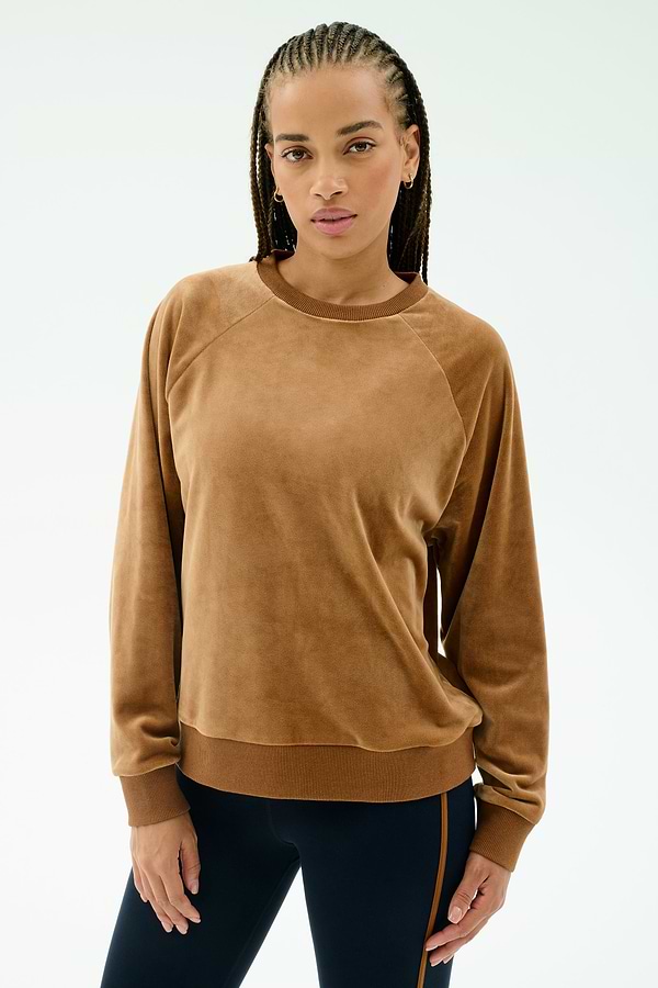 A woman with braided hair wears the SPLITS59 Joy Velour Crewneck in Honey, paired with dark side-striped stretch pants, standing against a plain light background.