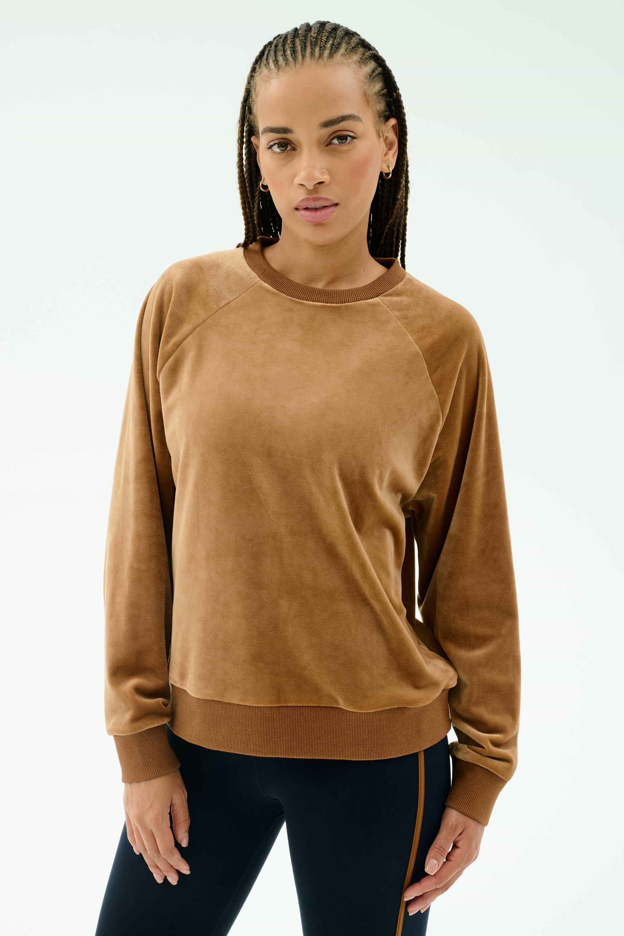 A woman with braided hair wears the SPLITS59 Joy Velour Crewneck in Honey, paired with dark side-striped stretch pants, standing against a plain light background.