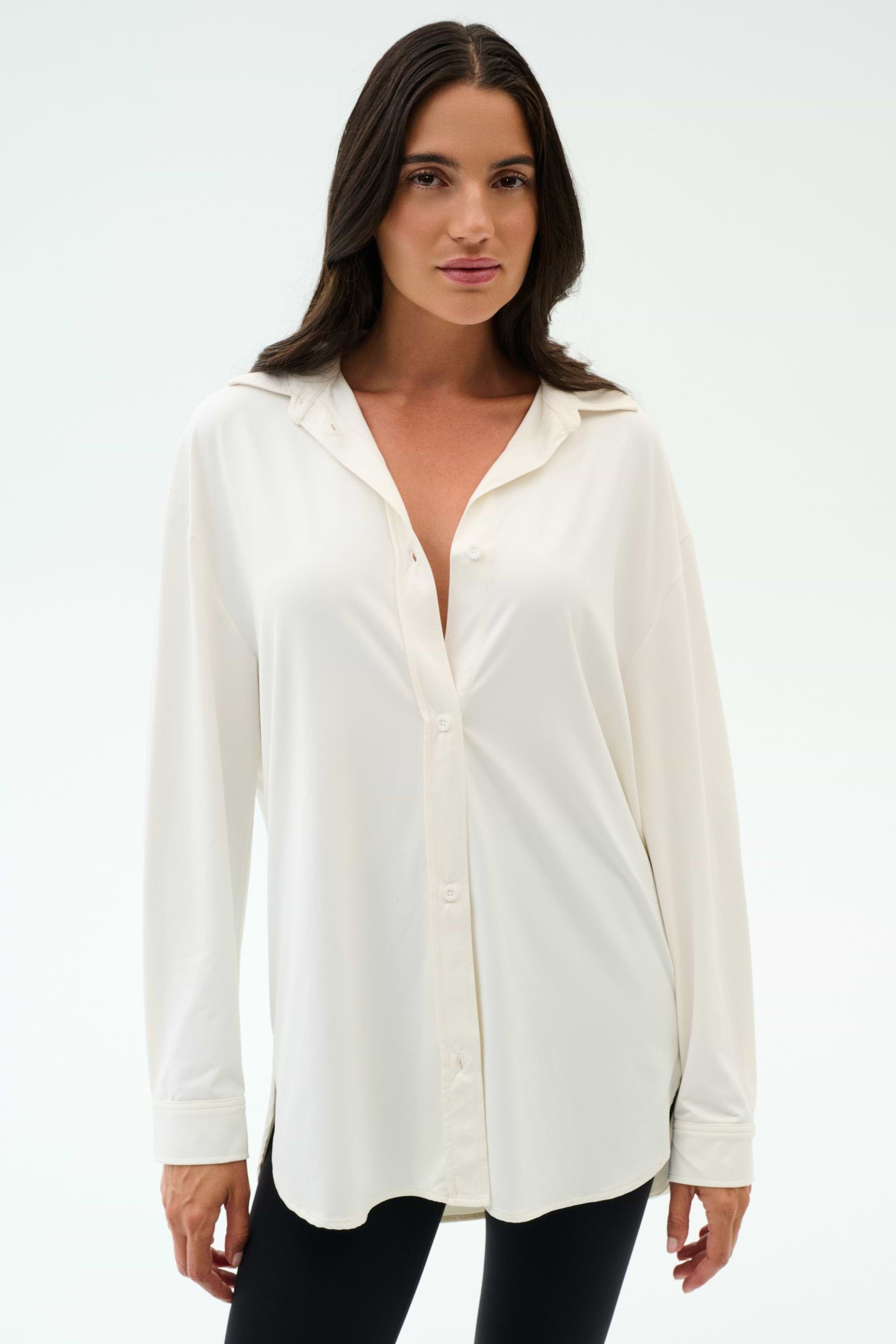 A woman with long brown hair models the SPLITS59 Kate Airweight Lite Button Down in white, paired with black pants, standing against a plain white background.
