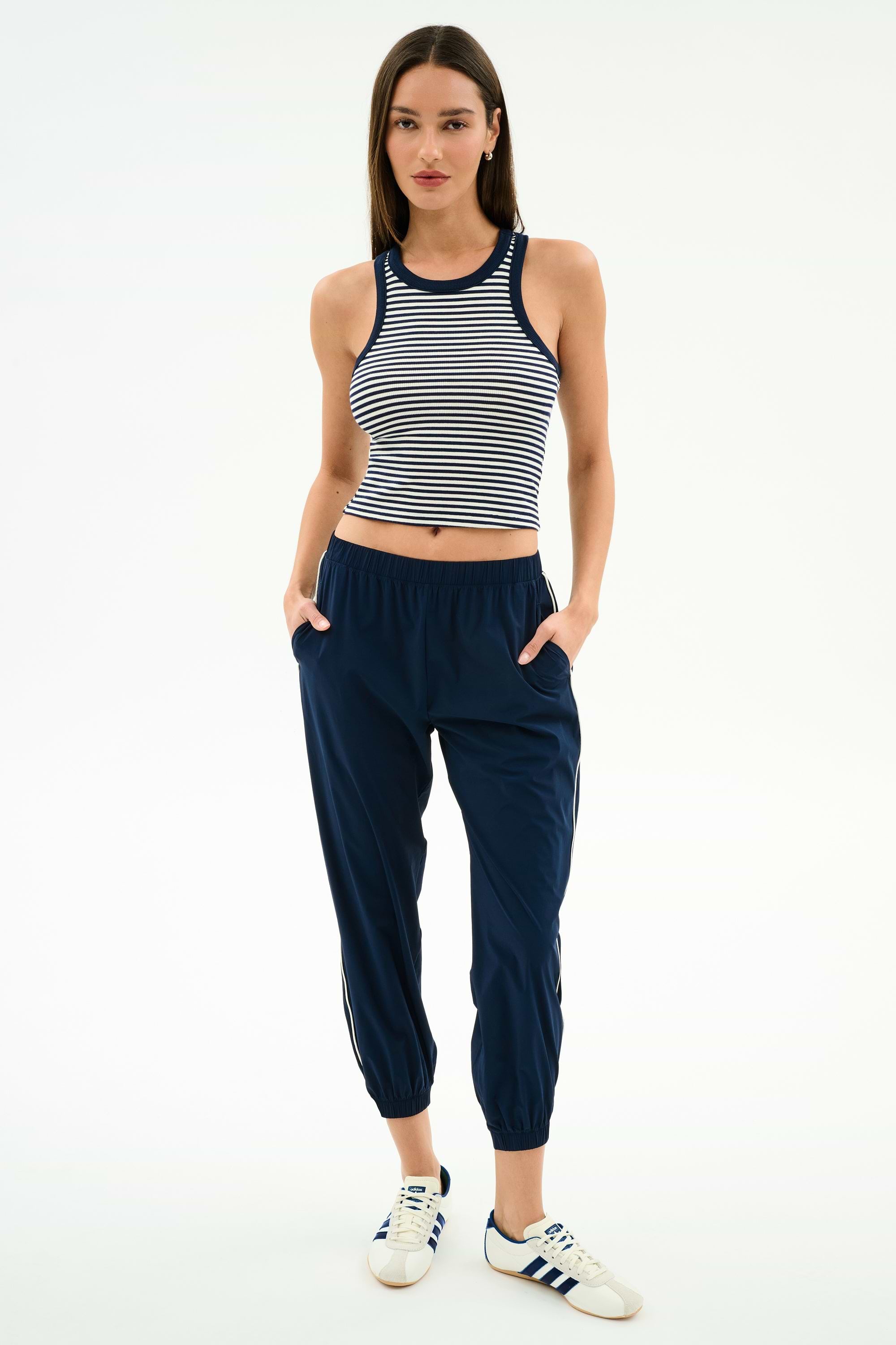 A woman poses with hands in pockets, wearing the SPLITS59 Kiki Rib Crop Tank in White/Indigo Stripe, navy jogger pants, and white Adidas sneakers against a plain white background.