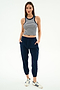 A woman poses with hands in pockets, wearing the SPLITS59 Kiki Rib Crop Tank in White/Indigo Stripe, navy jogger pants, and white Adidas sneakers against a plain white background.