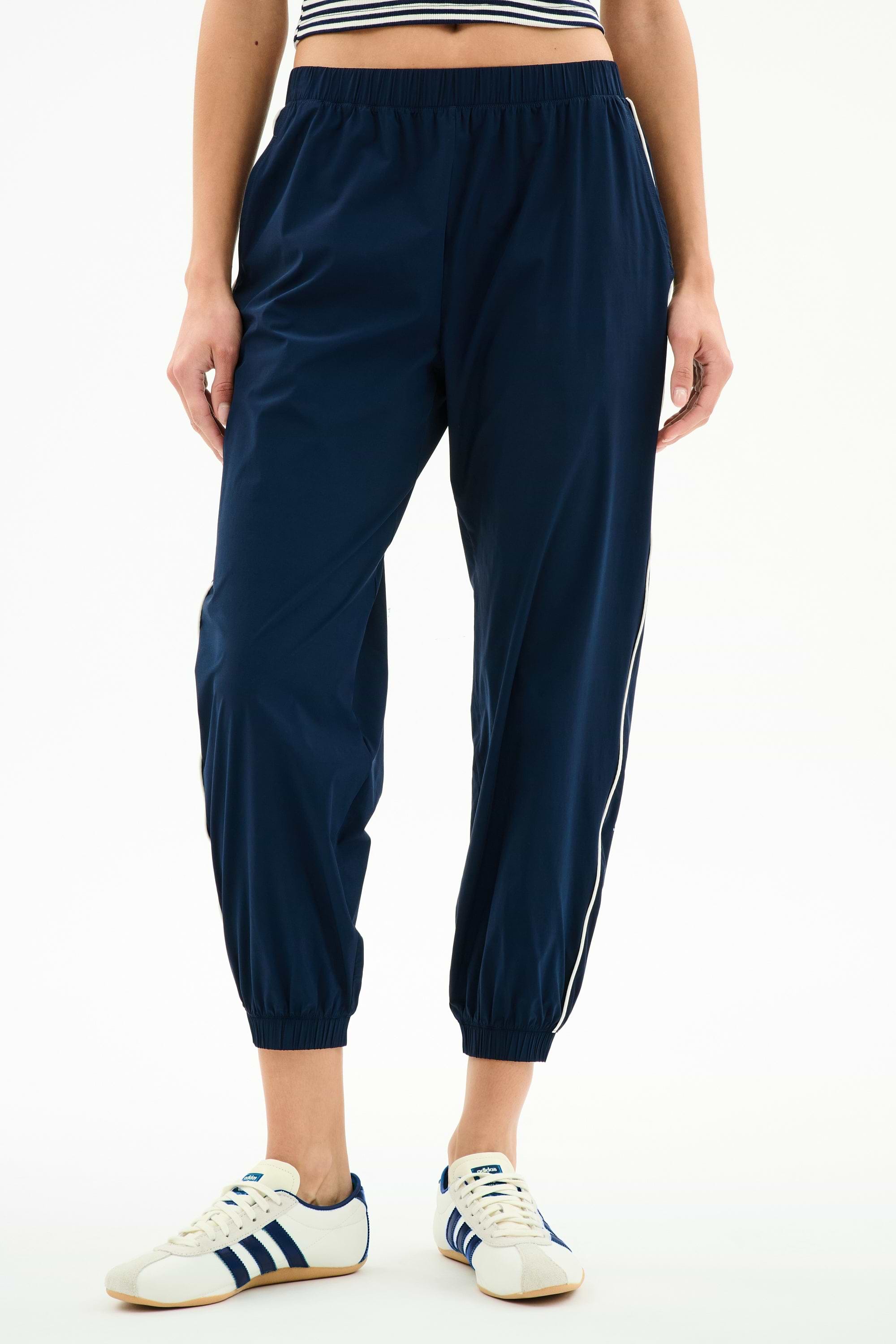Model wears SPLITS59 Megan Woven Nylon Jogger w/ Piping in Indigo/Creme, featuring a stretchy nylon-spandex blend, paired with white sneakers with blue stripes and a cropped striped top, shown from the waist down against a plain background.