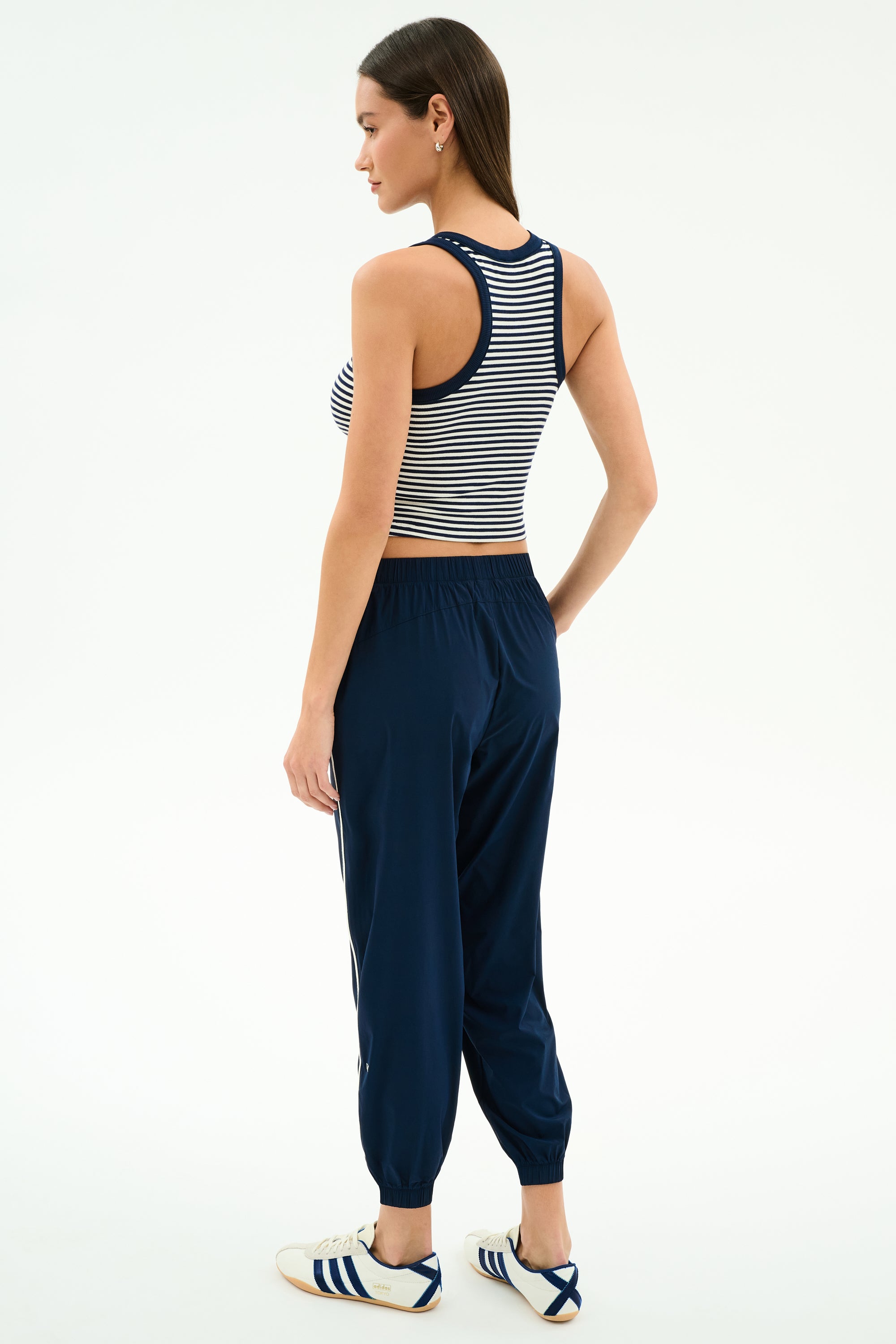 A woman stands against a white background, wearing the SPLITS59 Kiki Rib Crop Tank in White/Indigo Stripe, navy jogger pants, and white sneakers with blue stripes—an ideal gym look—while facing slightly away from the camera.