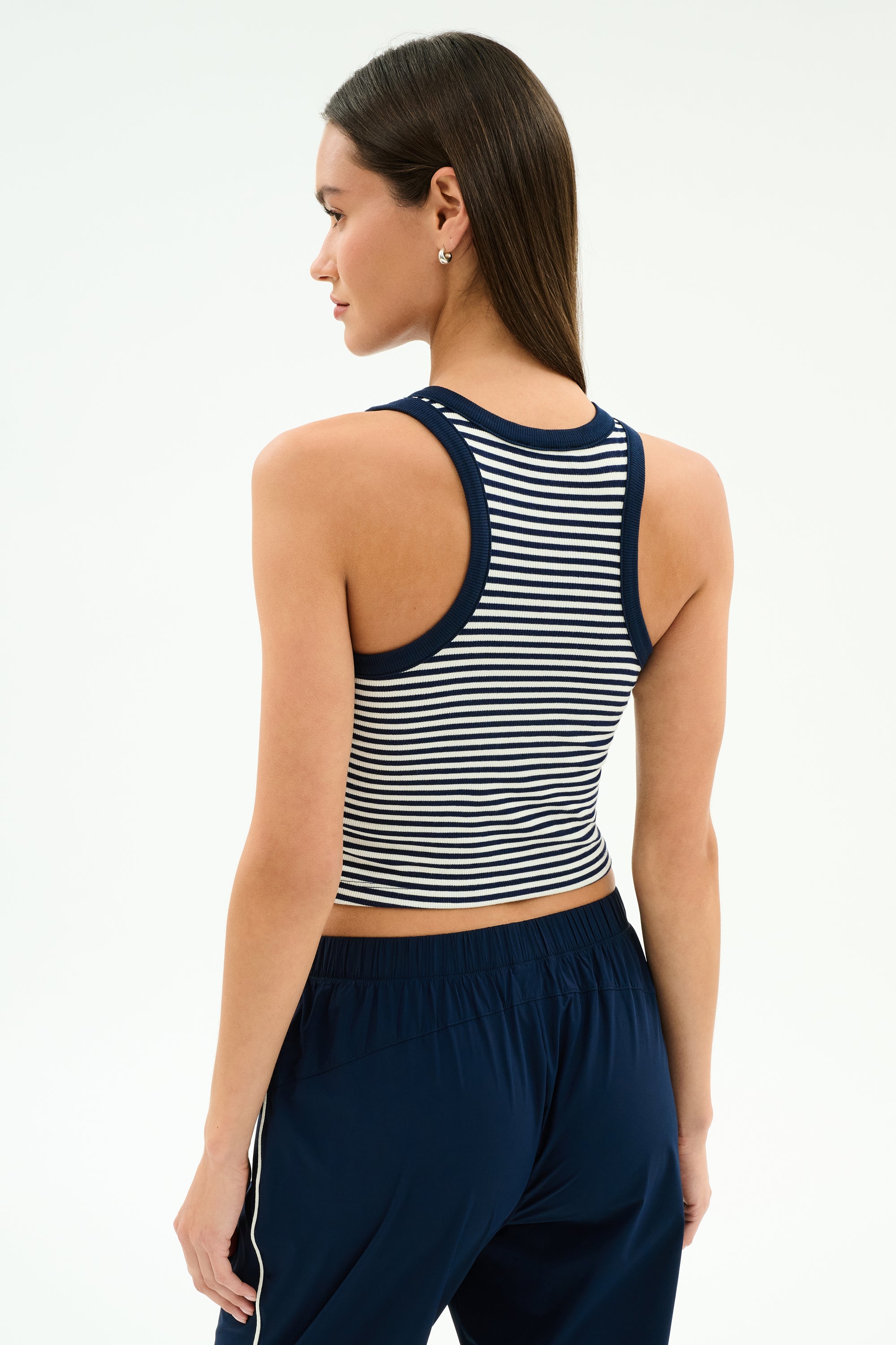 A woman faces away against a plain background, wearing the SPLITS59 Kiki Rib Crop Tank in White/Indigo Stripe with navy pants featuring white side stripes.