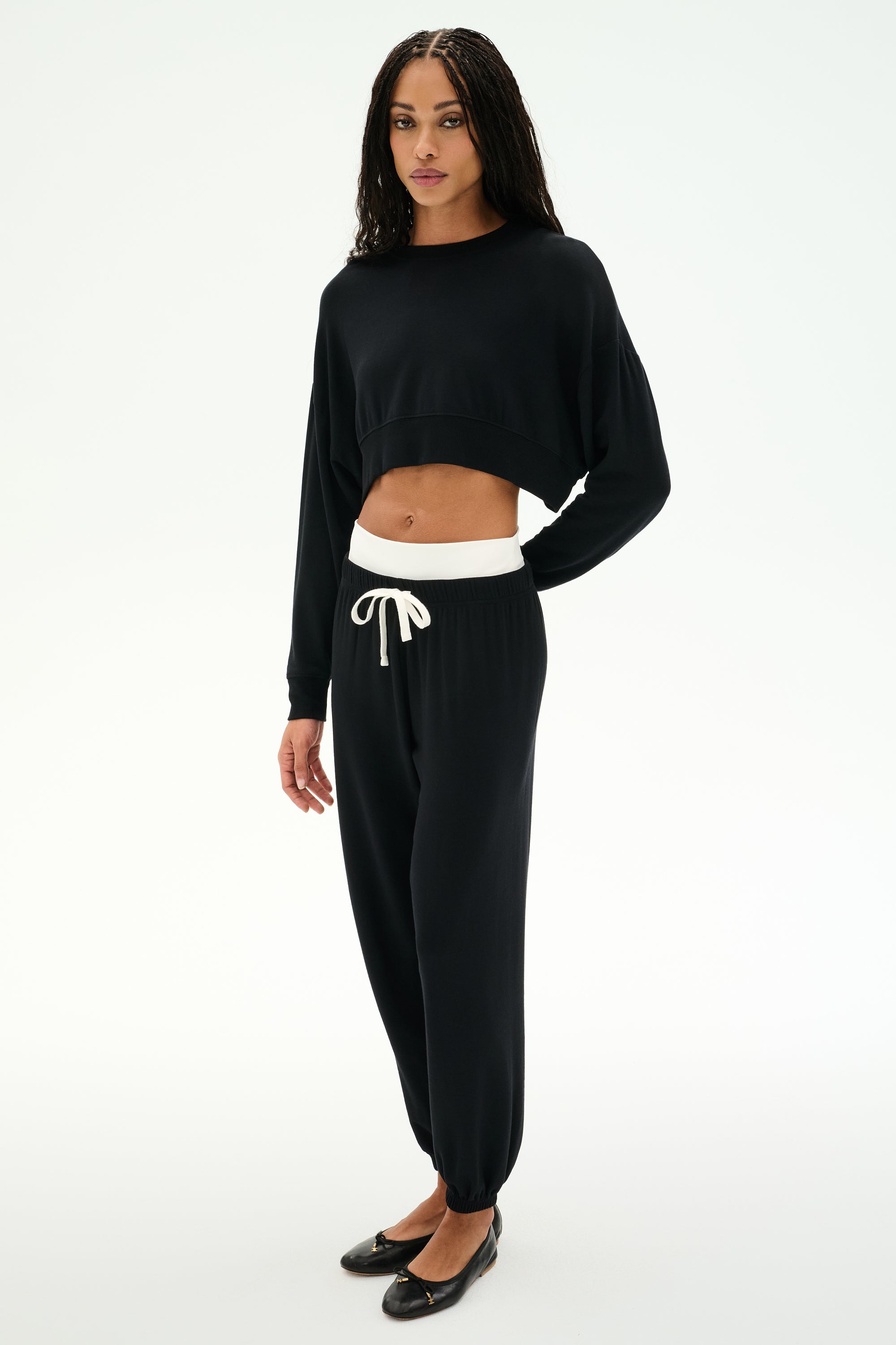 A person stands against a plain white background wearing the SPLITS59 Noah Fleece Crop Sweatshirt in black, styled with black drawstring jogger pants featuring a white waistband and paired with black flats.