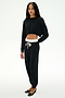 A person stands against a plain white background wearing the SPLITS59 Noah Fleece Crop Sweatshirt in black, styled with black drawstring jogger pants featuring a white waistband and paired with black flats.