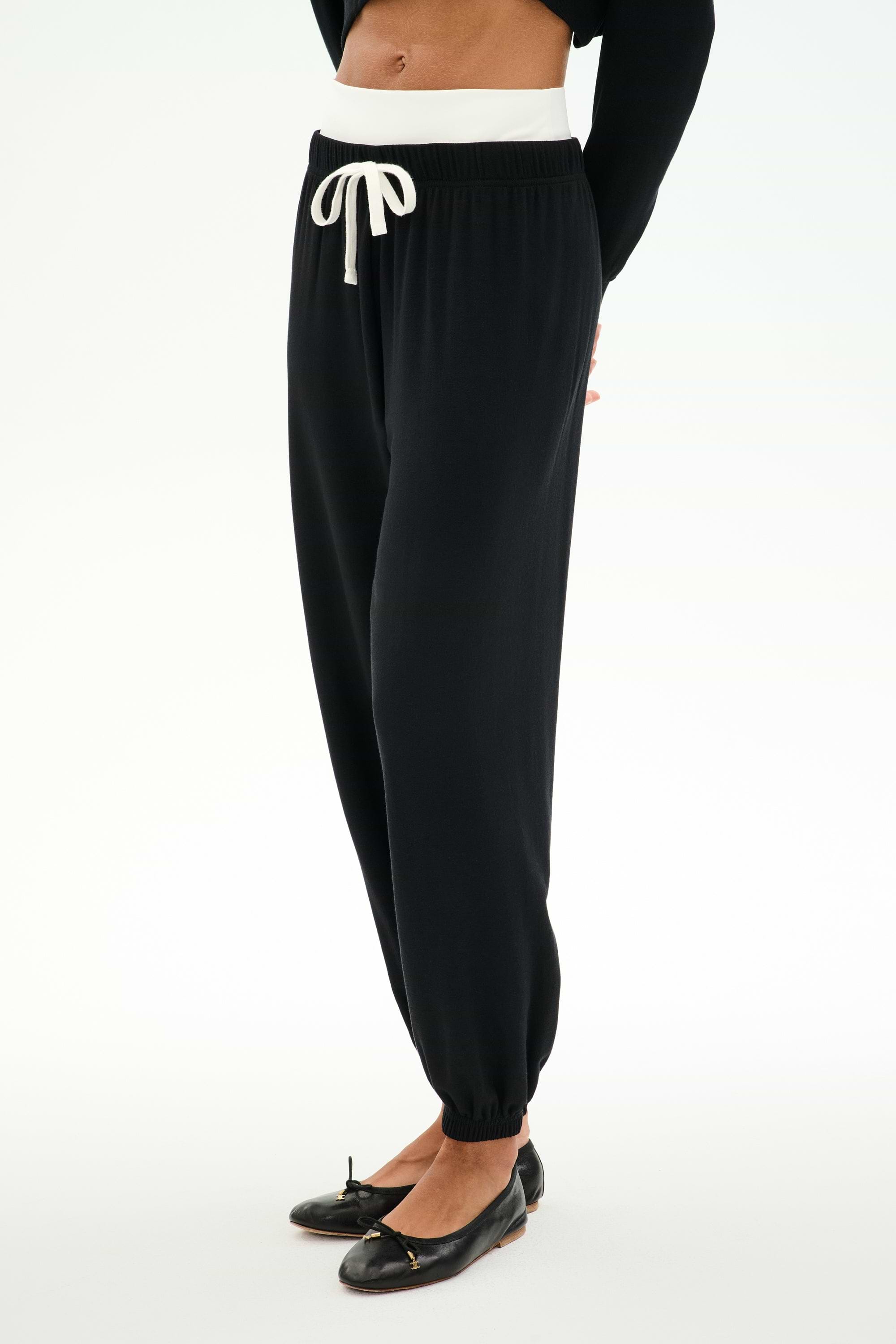 A person wears SPLITS59 Andie Dual Waist Sweatpant in Black/White, featuring a white drawstring and waistband, soft modal blend fabric, paired with a black long-sleeve top and ballet flats, standing against a white background.