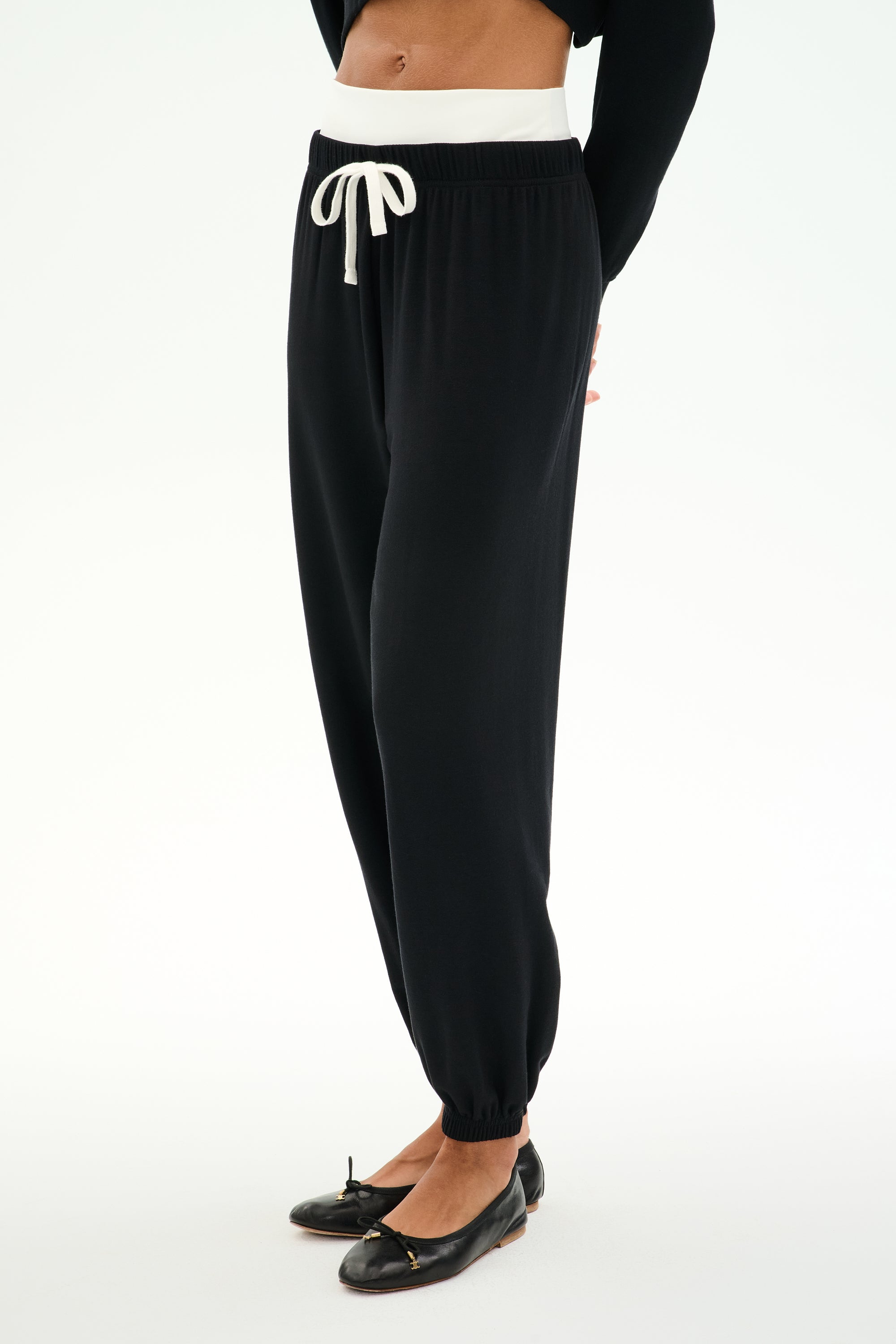 A person wears SPLITS59 Andie Dual Waist Sweatpant in Black/White, featuring a white drawstring and waistband, soft modal blend fabric, paired with a black long-sleeve top and ballet flats, standing against a white background.