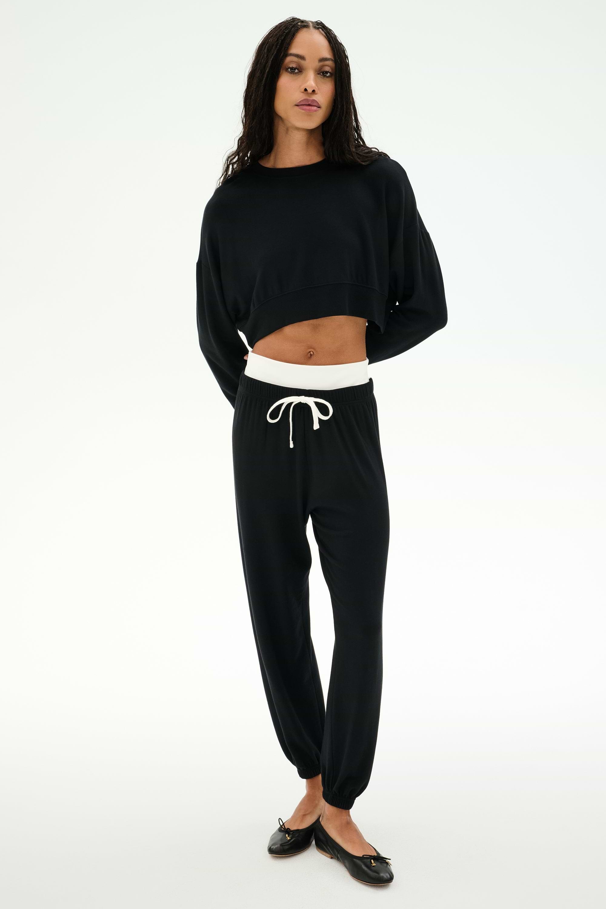 A woman stands relaxed wearing the SPLITS59 Noah Fleece Crop Sweatshirt in black, paired with black drawstring joggers featuring a white waistband and black flats, set against a plain white background.