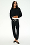 A woman stands relaxed wearing the SPLITS59 Noah Fleece Crop Sweatshirt in black, paired with black drawstring joggers featuring a white waistband and black flats, set against a plain white background.