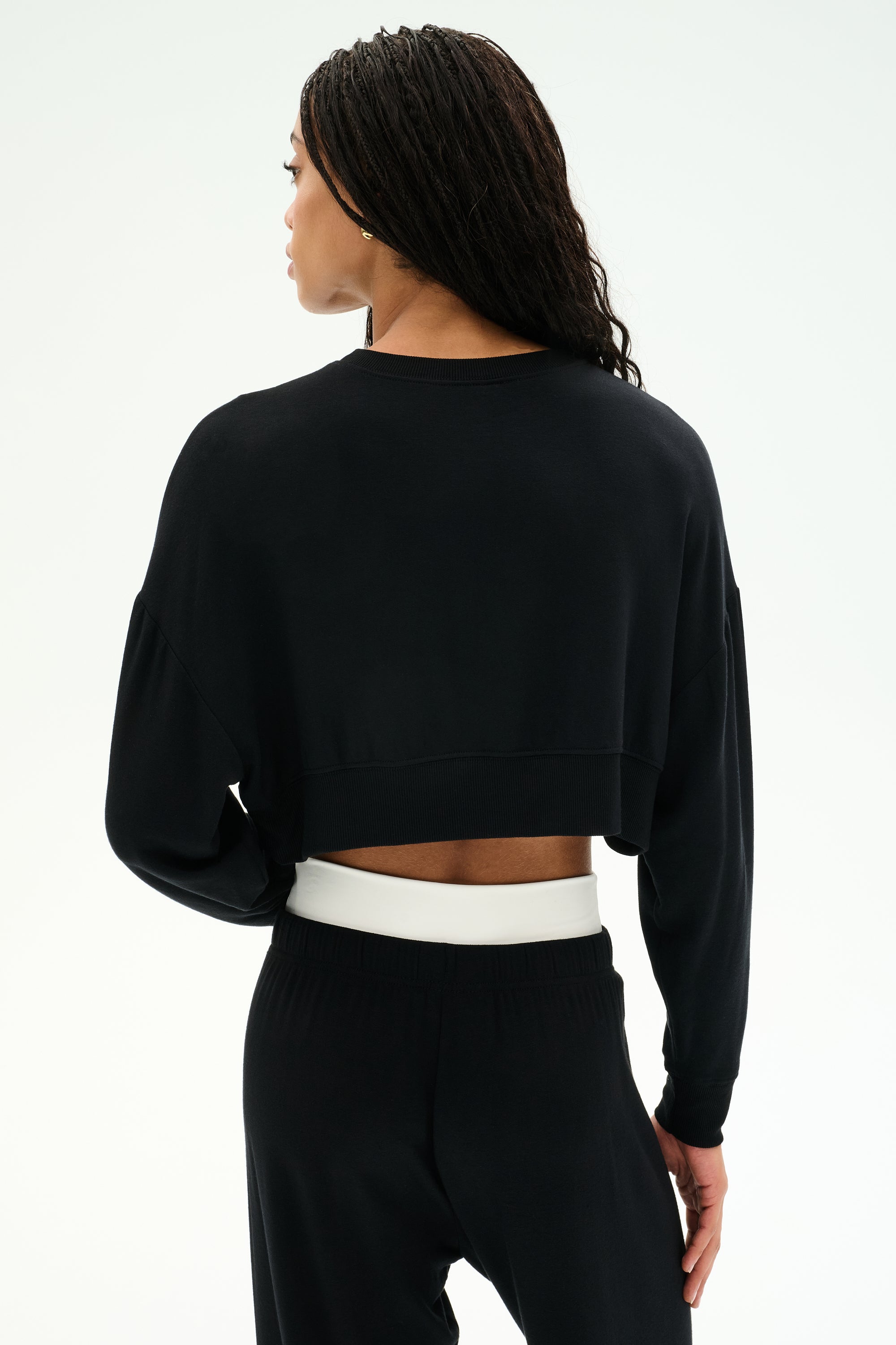 A person with long braided hair faces away from the camera, wearing the SPLITS59 Noah Fleece Crop Sweatshirt in black and matching black pants against a plain white background.