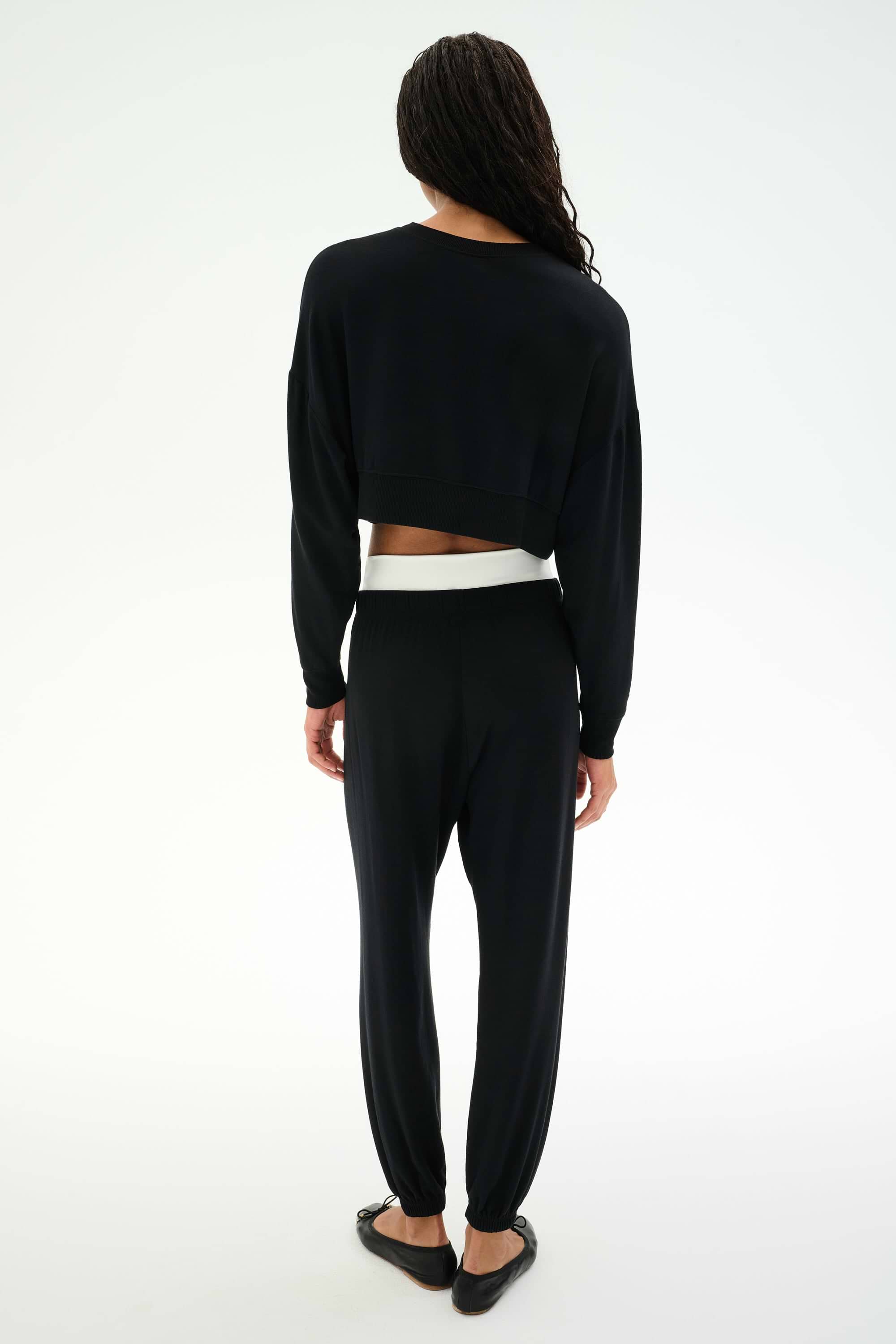 A person with long hair faces away, wearing the SPLITS59 Noah Fleece Crop Sweatshirt in black, paired with black pants and flats, set against a plain white background.