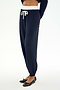 A person is wearing the SPLITS59 Andie Dual Waist Sweatpant in Indigo/White, featuring a white drawstring, paired with black ballet flats and a cropped top made from ultra-luxe fleece. The background is plain and light-colored.