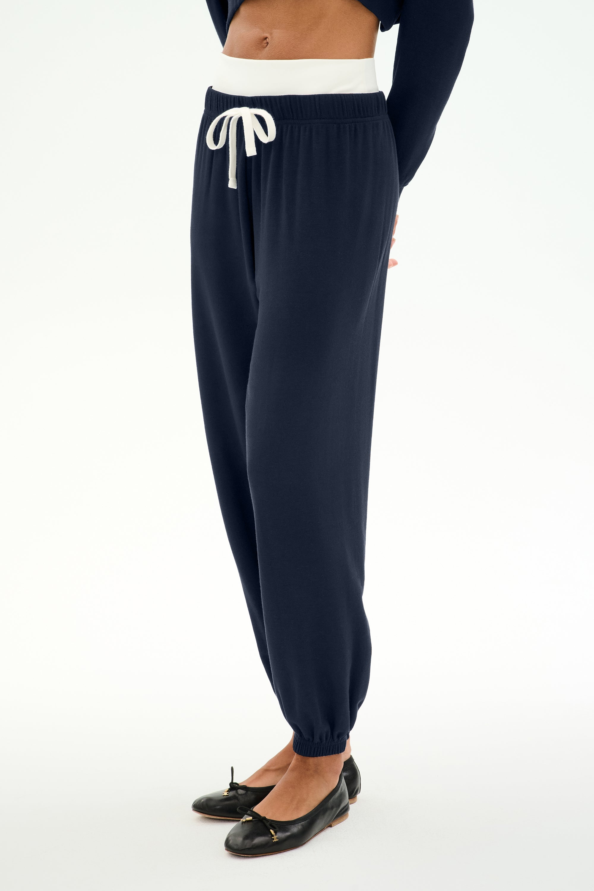 A person is wearing the SPLITS59 Andie Dual Waist Sweatpant in Indigo/White, featuring a white drawstring, paired with black ballet flats and a cropped top made from ultra-luxe fleece. The background is plain and light-colored.