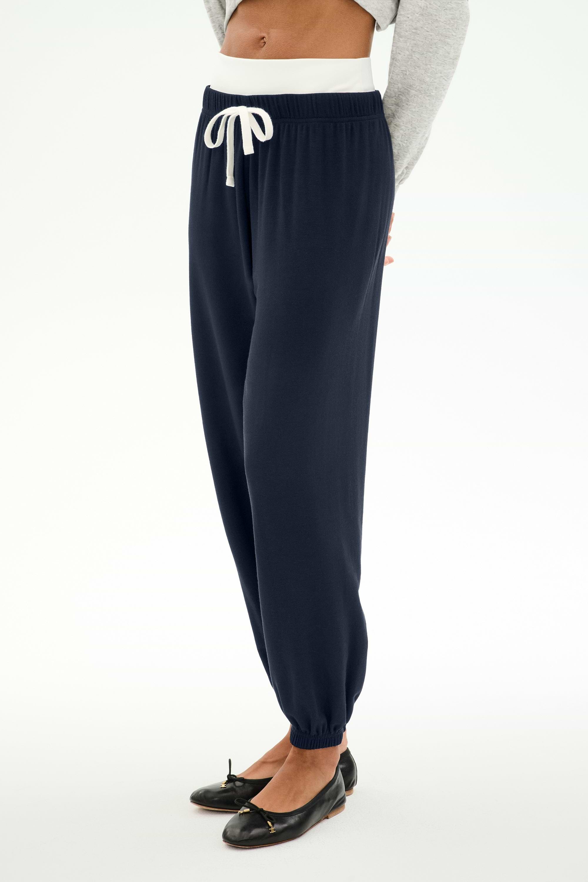 A person is wearing the SPLITS59 Andie Dual Waist Sweatpant in Indigo/White, featuring a comfortable baggy sweatpant fit and a white drawstring. The joggers are styled with a gray sweater and black ballet flats, against a white background. Made in Los Angeles.