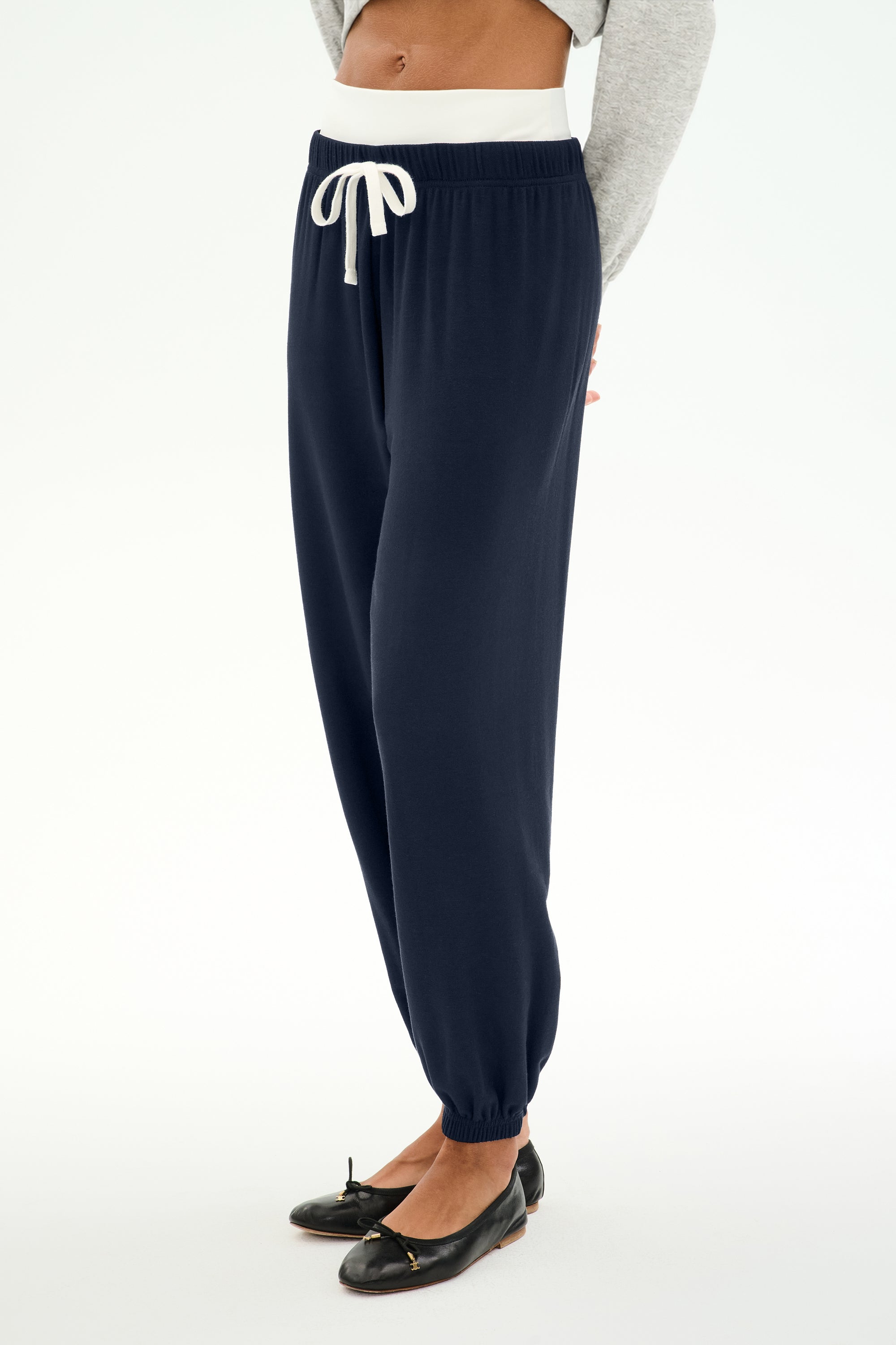 A person is wearing the SPLITS59 Andie Dual Waist Sweatpant in Indigo/White, featuring a comfortable baggy sweatpant fit and a white drawstring. The joggers are styled with a gray sweater and black ballet flats, against a white background. Made in Los Angeles.