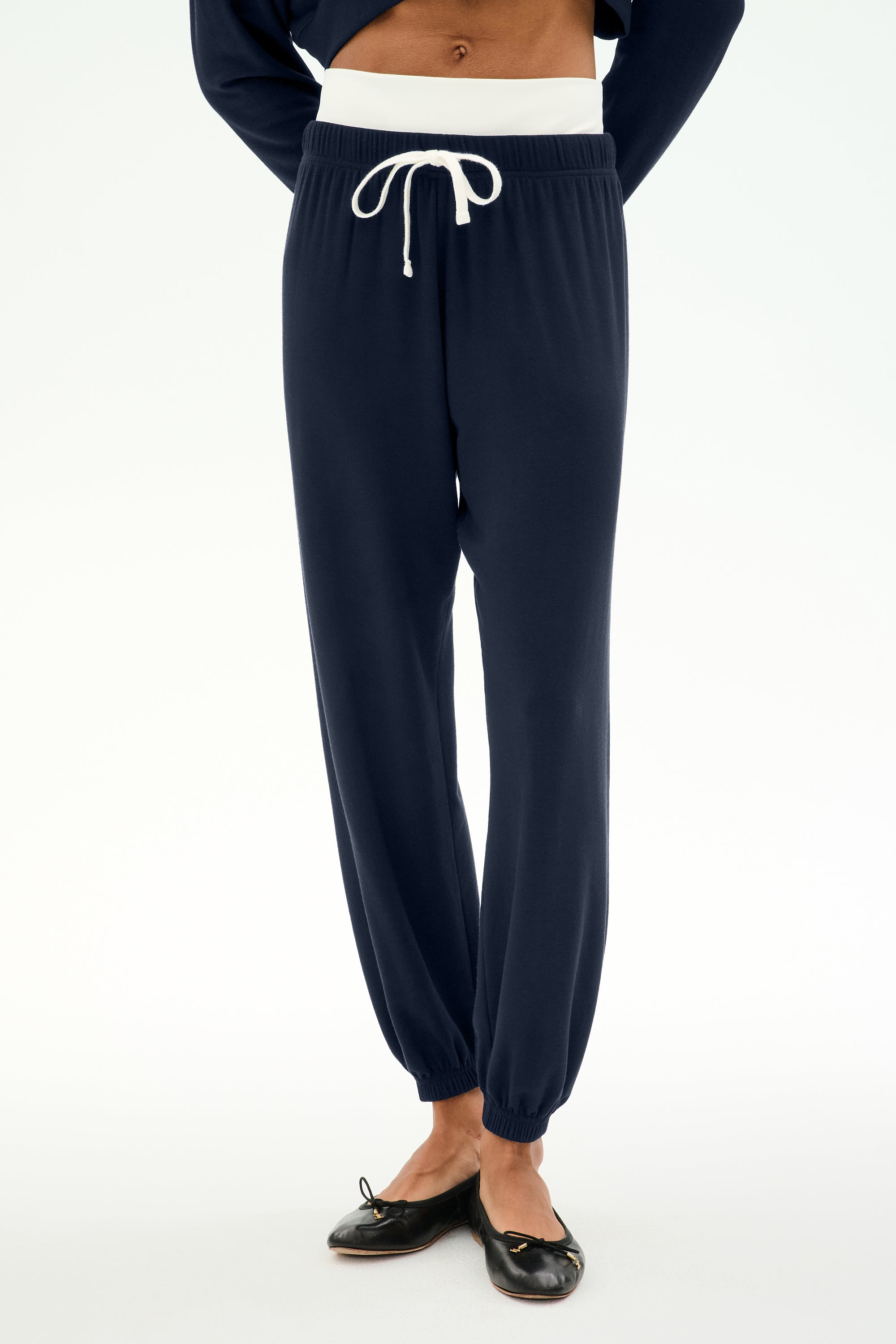 A model wears the SPLITS59 Andie Dual Waist Sweatpant in Indigo/White, crafted from ultra-luxe fleece with a contrasting white drawstring and white waistband. Styled with black ballet flats and photographed against a plain white background, these sweatpants are proudly made in Los Angeles.