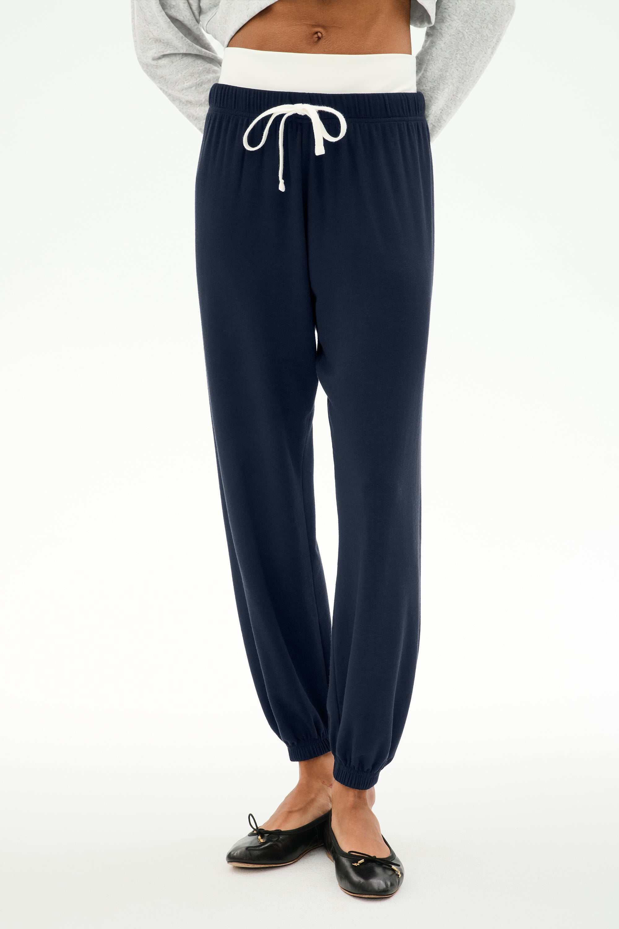 A person wears the SPLITS59 Andie Dual Waist Sweatpant in Indigo/White, featuring a relaxed fit with a white drawstring, paired with a gray top and black ballet flats, standing against a white background. These fleece sweatpants are crafted in Los Angeles to offer effortless comfort and style.