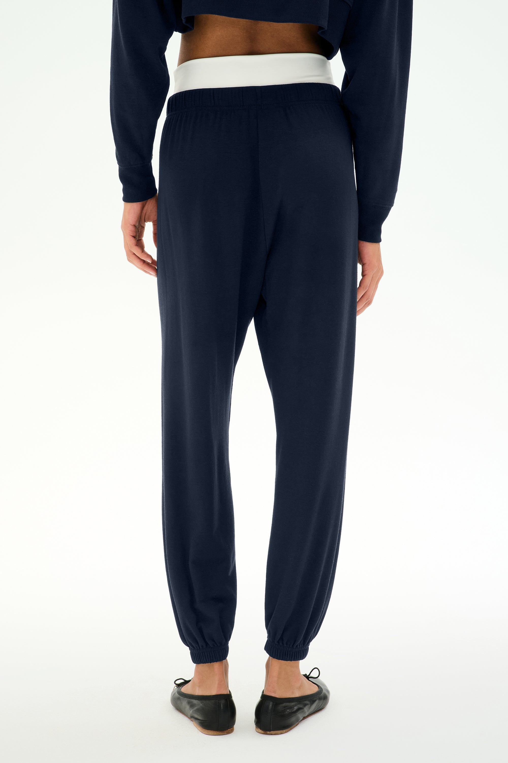 A person stands with their back to the camera on a plain white background, wearing the SPLITS59 Andie Dual Waist Sweatpant in Indigo/White, crafted from ultra-luxe fleece with an exposed white waistband, paired with black ballet flats.