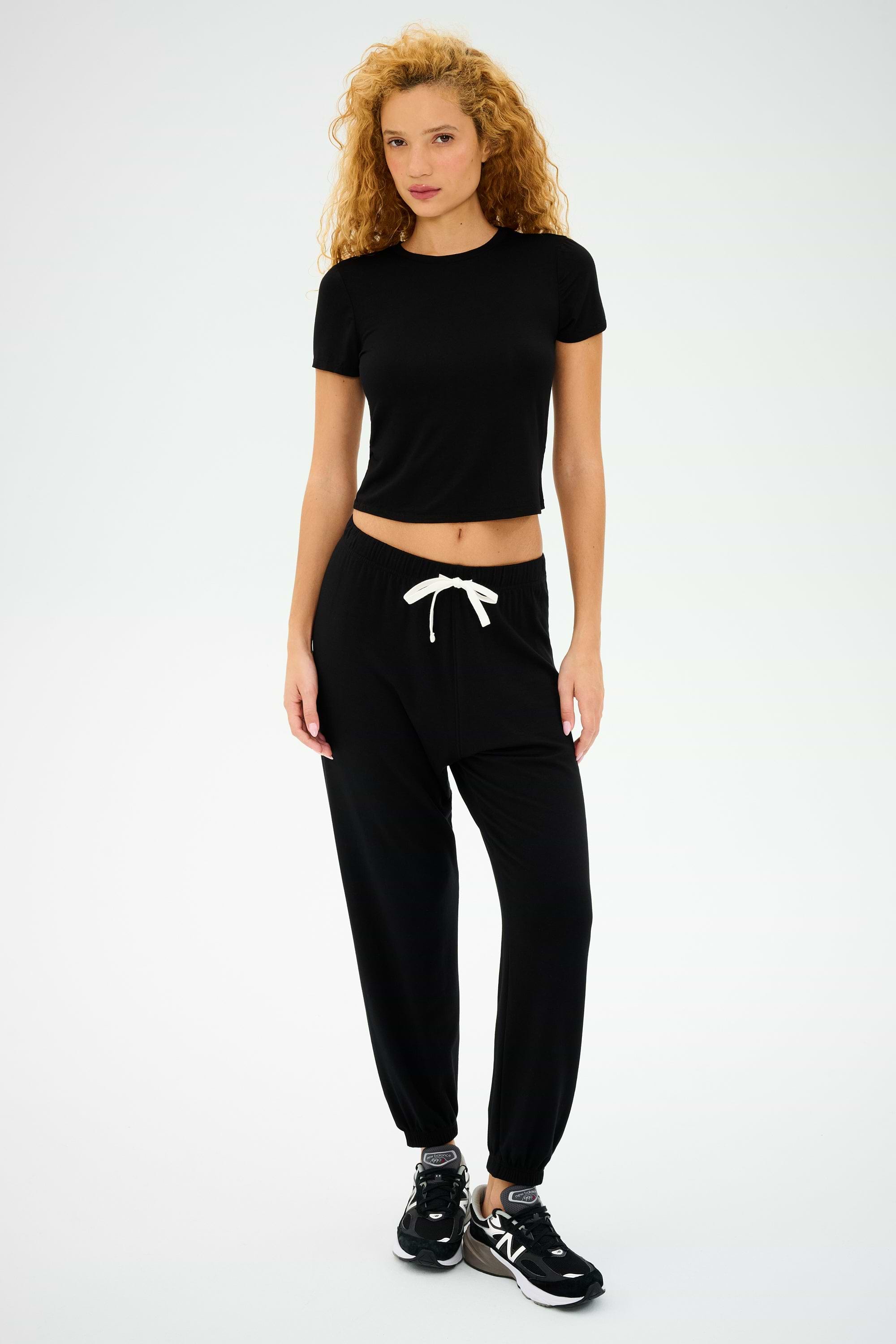 With curly hair, a person wears SPLITS59's Andie Oversized Fleece Sweatpant in black and a simple black t-shirt, matched with sleek sneakers against a plain backdrop. This stylish outfit, ideal for cool weather workouts, is proudly made in Los Angeles.