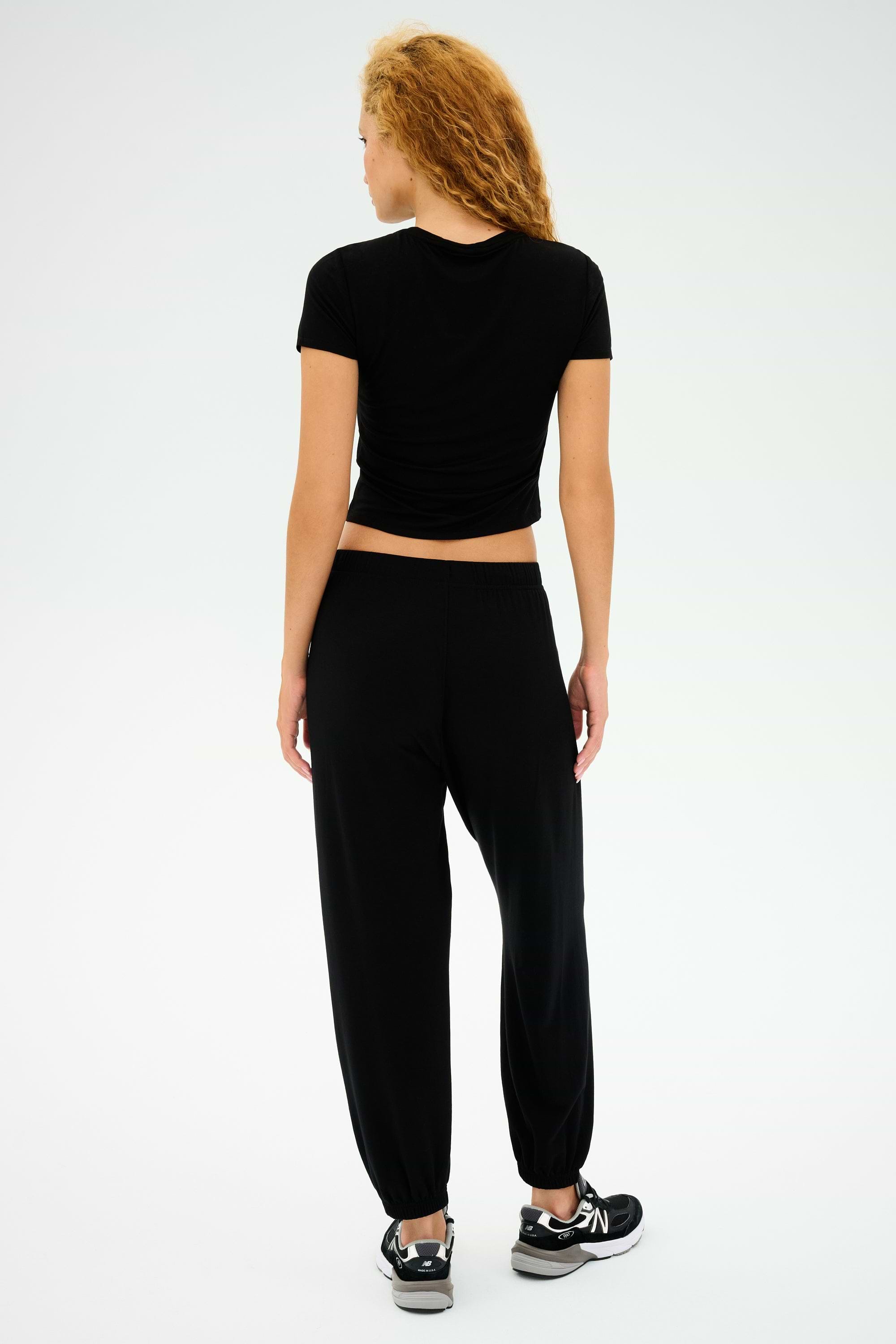A curly-haired person in a black t-shirt, wearing SPLITS59's Andie Oversized Fleece Sweatpants and black sneakers, stands against a light gray background. Ideal for cool weather workouts.