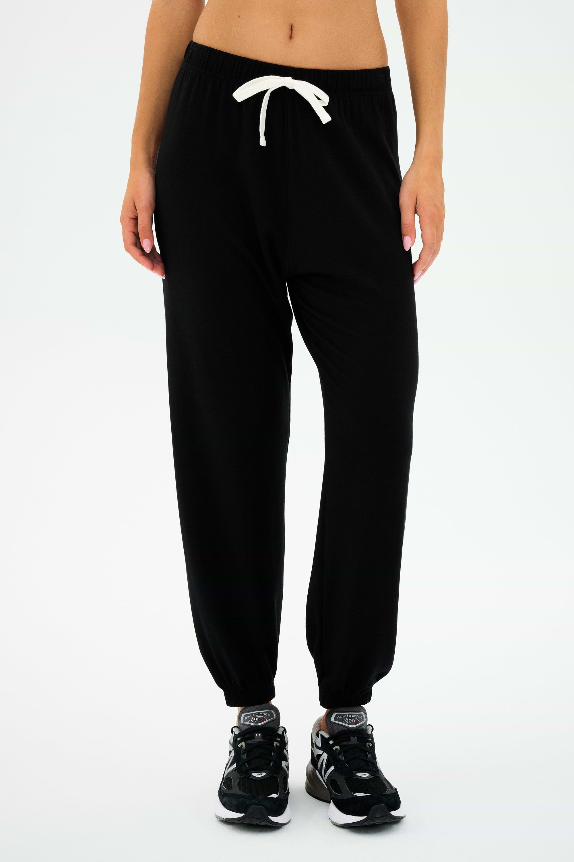 A person donning the SPLITS59 Andie Oversized Fleece Sweatpant in Black, paired with black sneakers and a white crop top, stands against a plain white background. Ideal for cool weather workouts and proudly MADE IN LOS ANGELES.