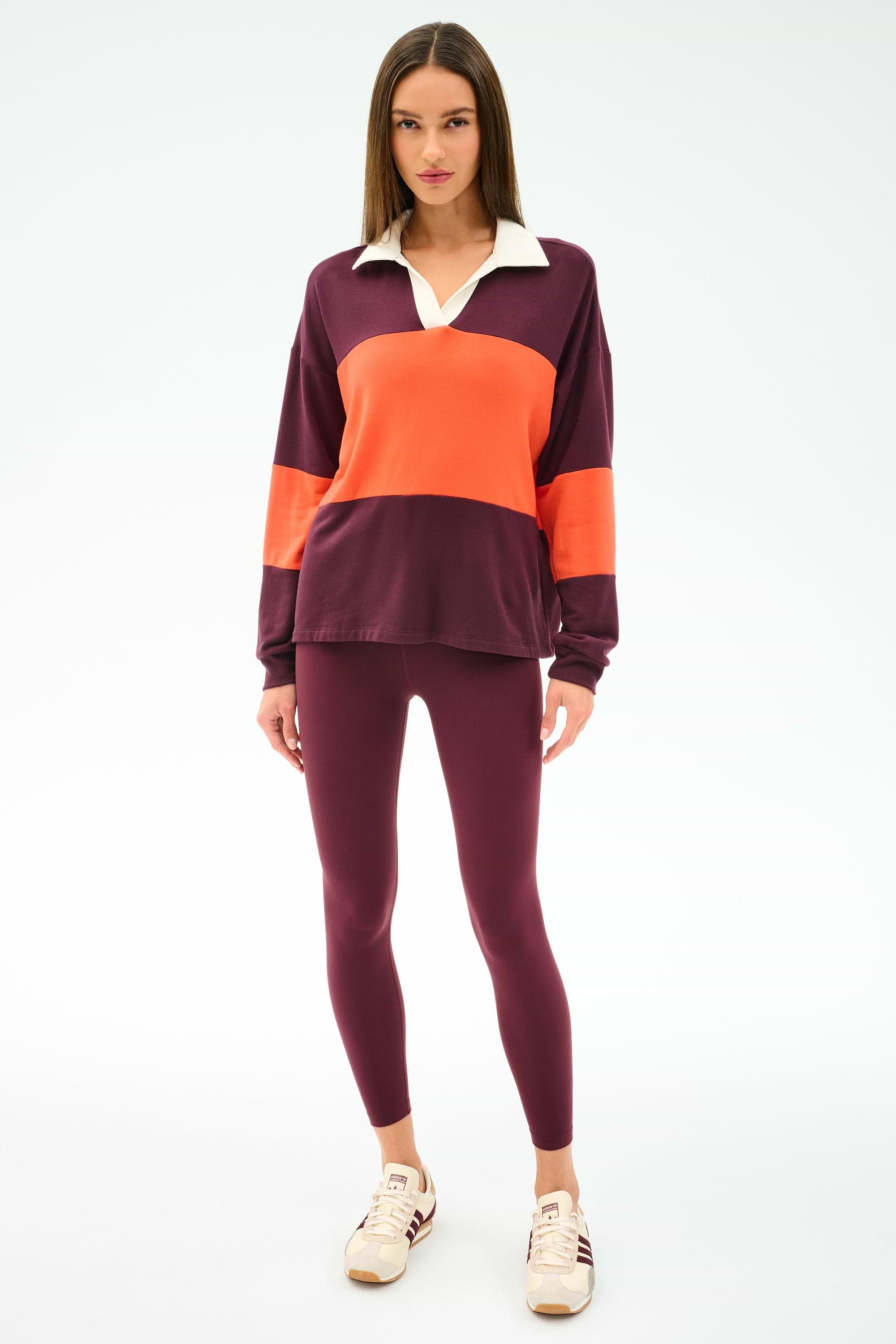 A woman stands against a plain background in the SPLITS59 Katie Fleece Rugby in Dark Cherry/Valencia, paired with maroon leggings and white sneakers.