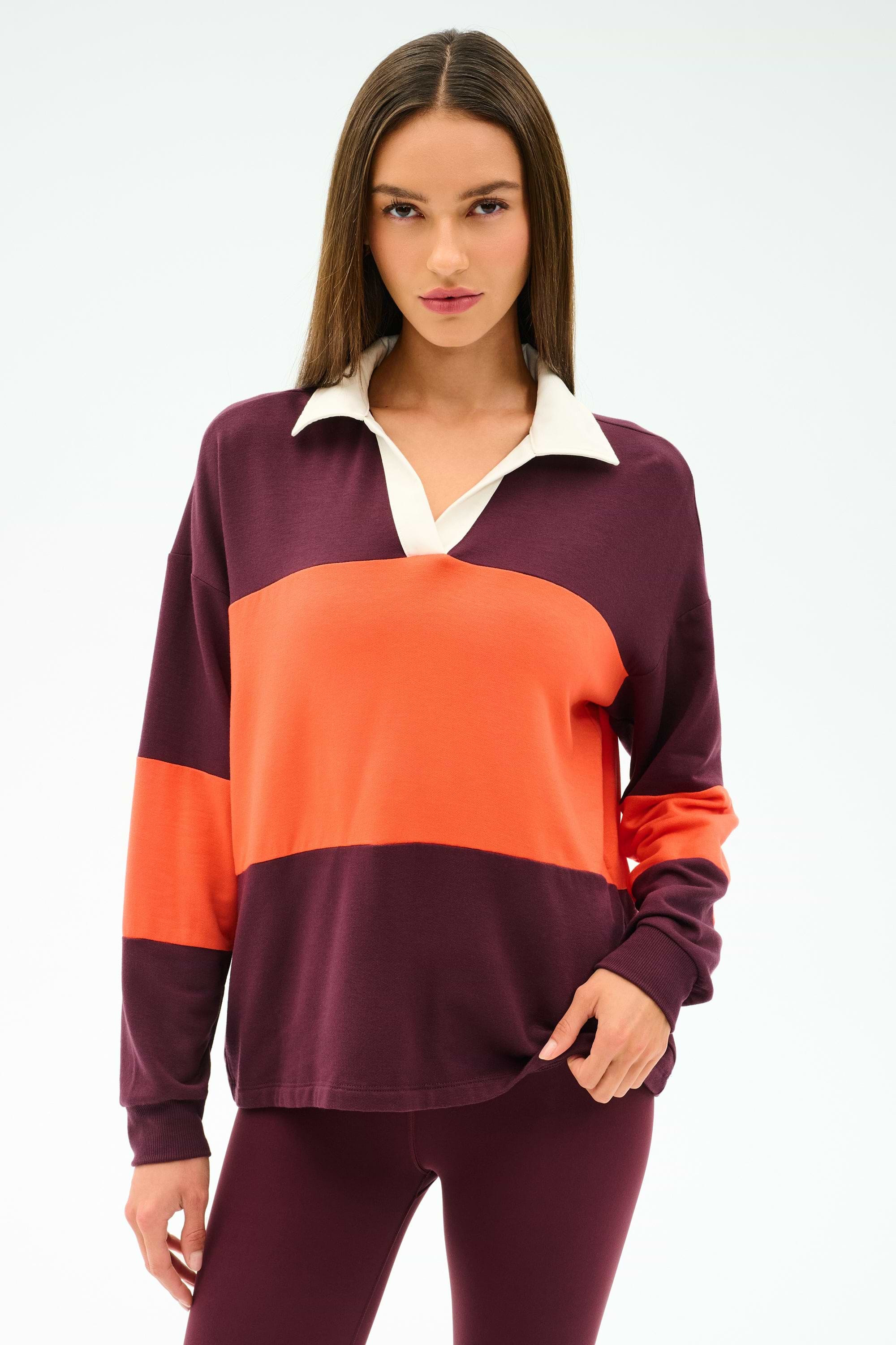 A woman with straight brown hair wears the SPLITS59 Katie Fleece Rugby in Dark Cherry/Valencia, paired with burgundy pants, standing against a plain light background.