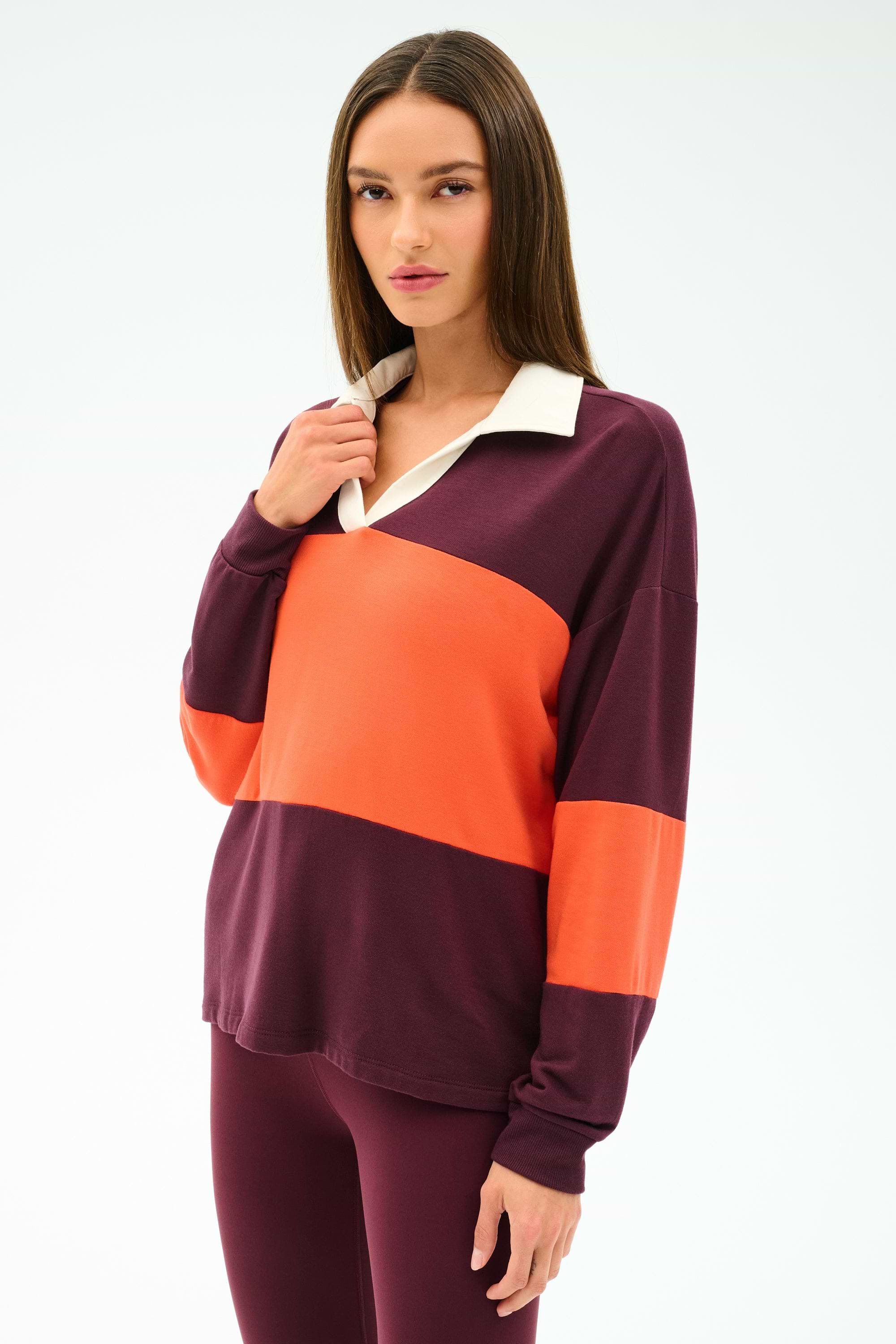 A woman poses against a plain background wearing the SPLITS59 Katie Fleece Rugby in Dark Cherry/Valencia— a long-sleeve maroon and orange color-blocked top—paired with matching maroon pants.