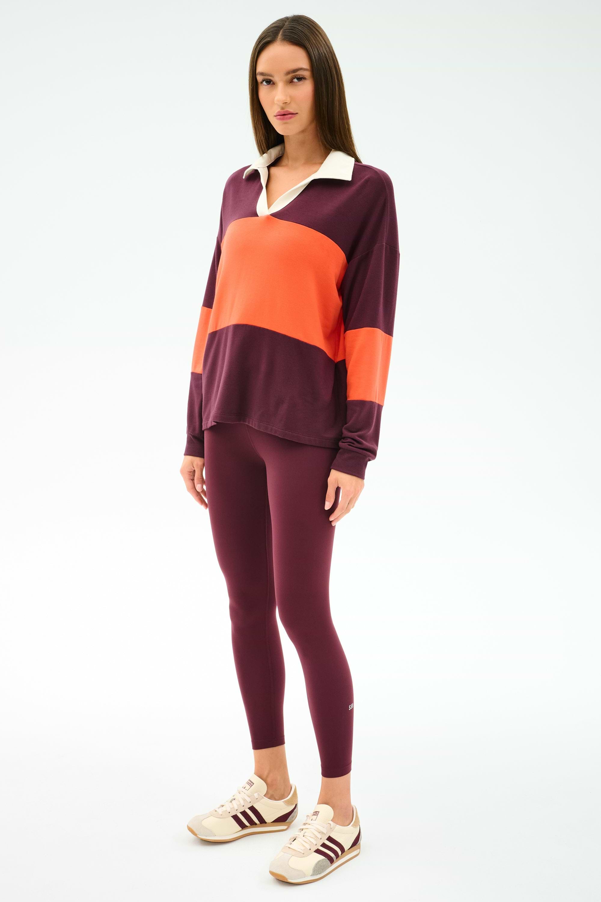 A woman stands against a plain white background wearing the SPLITS59 Katie Fleece Rugby in Dark Cherry/Valencia, maroon leggings, and beige sneakers with maroon stripes.