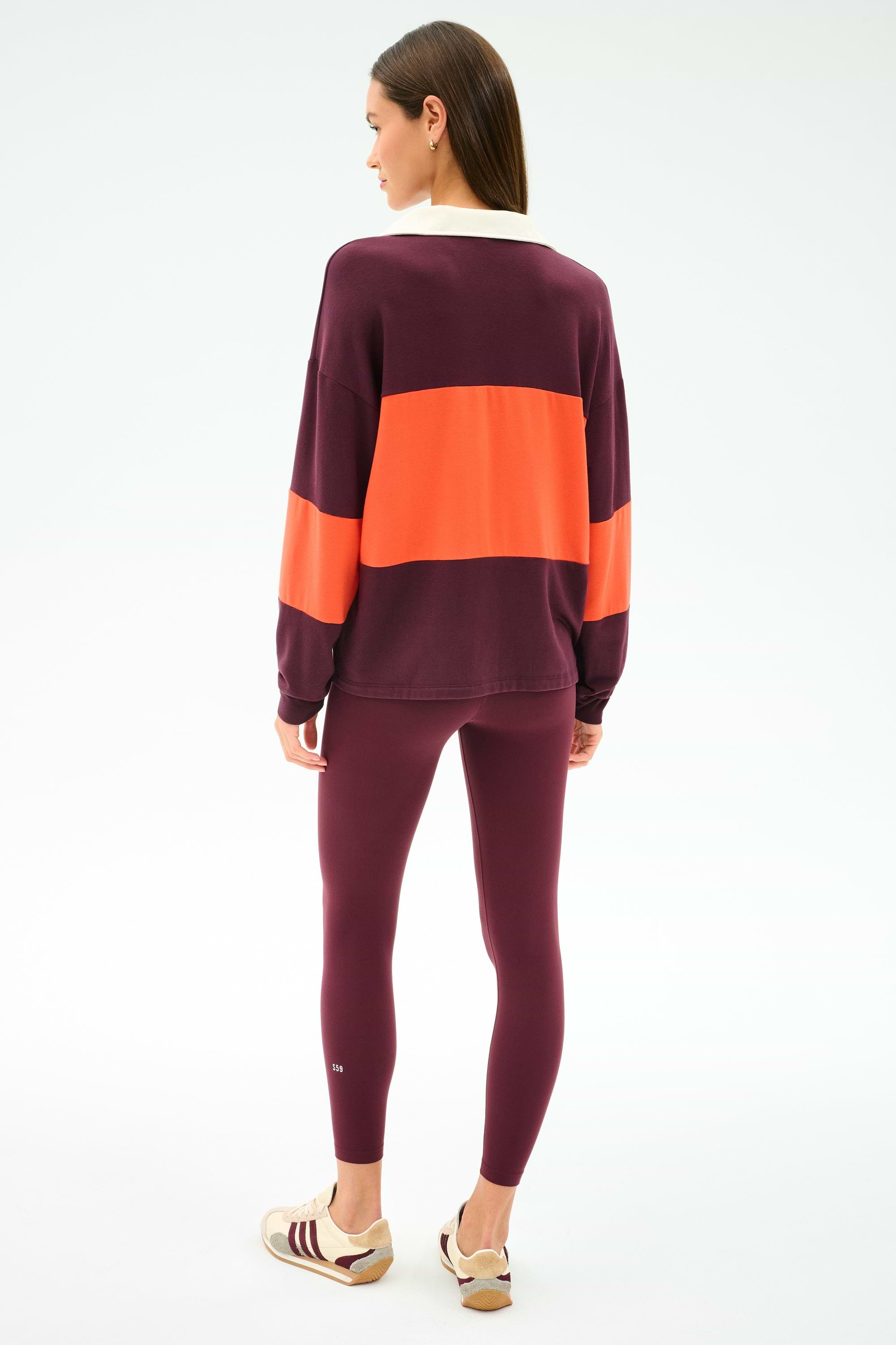 A woman stands facing away, wearing the SPLITS59 Katie Fleece Rugby in Dark Cherry/Valencia with matching maroon spandex leggings and beige sneakers featuring maroon stripes.