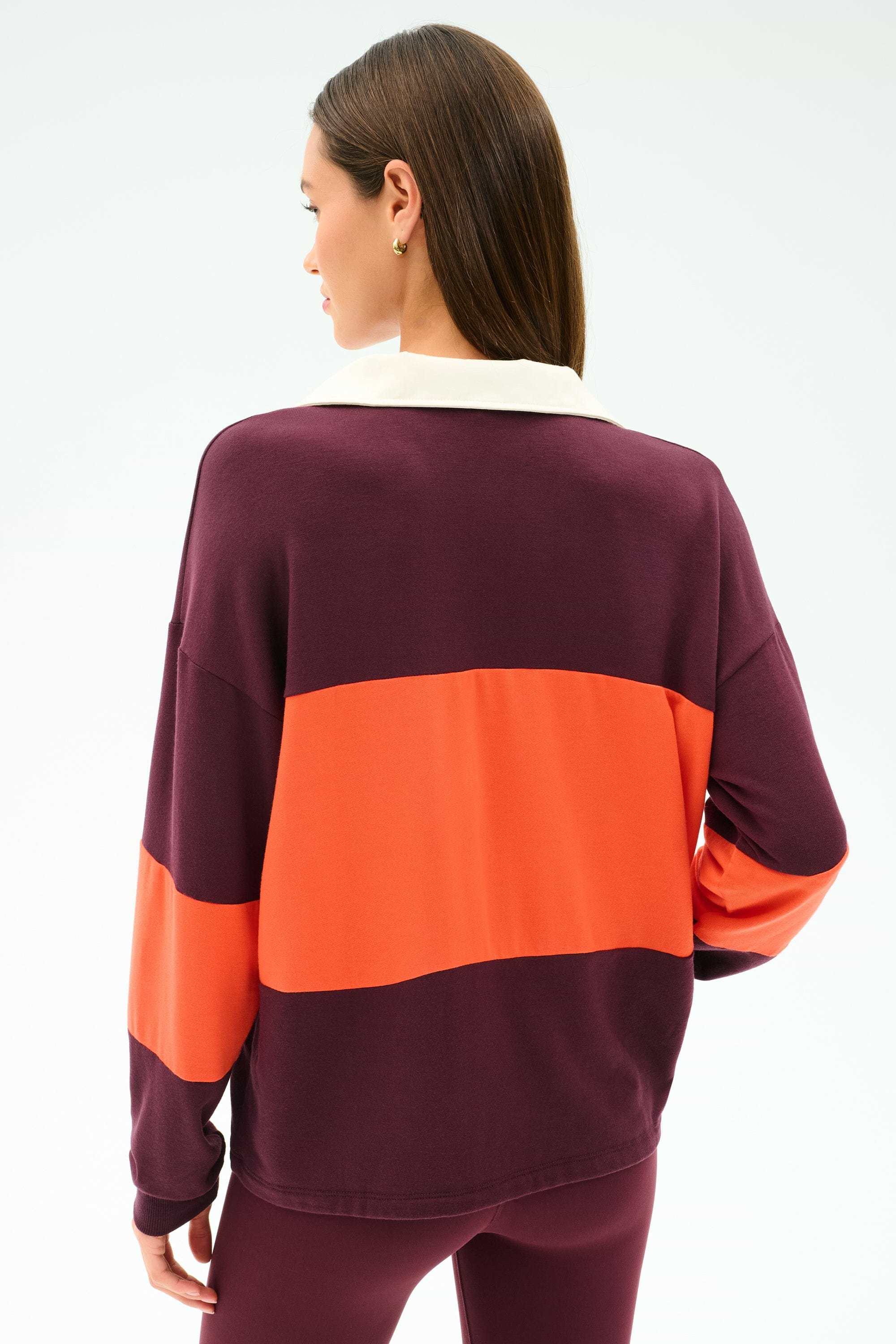 A woman faces away, wearing the SPLITS59 Katie Fleece Rugby in Dark Cherry/Valencia—a long-sleeve top with wide maroon and orange stripes—paired with maroon modal-spandex leggings.