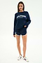 A woman wears the SPLITS59 Andie Oversized Fleece Varsity Graphic Sweatshirt in Indigo with "SPLITS59 Los Angeles" text, matching blue shorts with white trim, and white sneakers on a plain white background.
