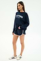 A woman stands against a plain background wearing the SPLITS59 Andie Oversized Fleece Varsity Graphic Sweatshirt in Indigo, featuring "Splits59 Los Angeles" on the front, paired with navy athletic shorts and white sneakers with navy stripes.