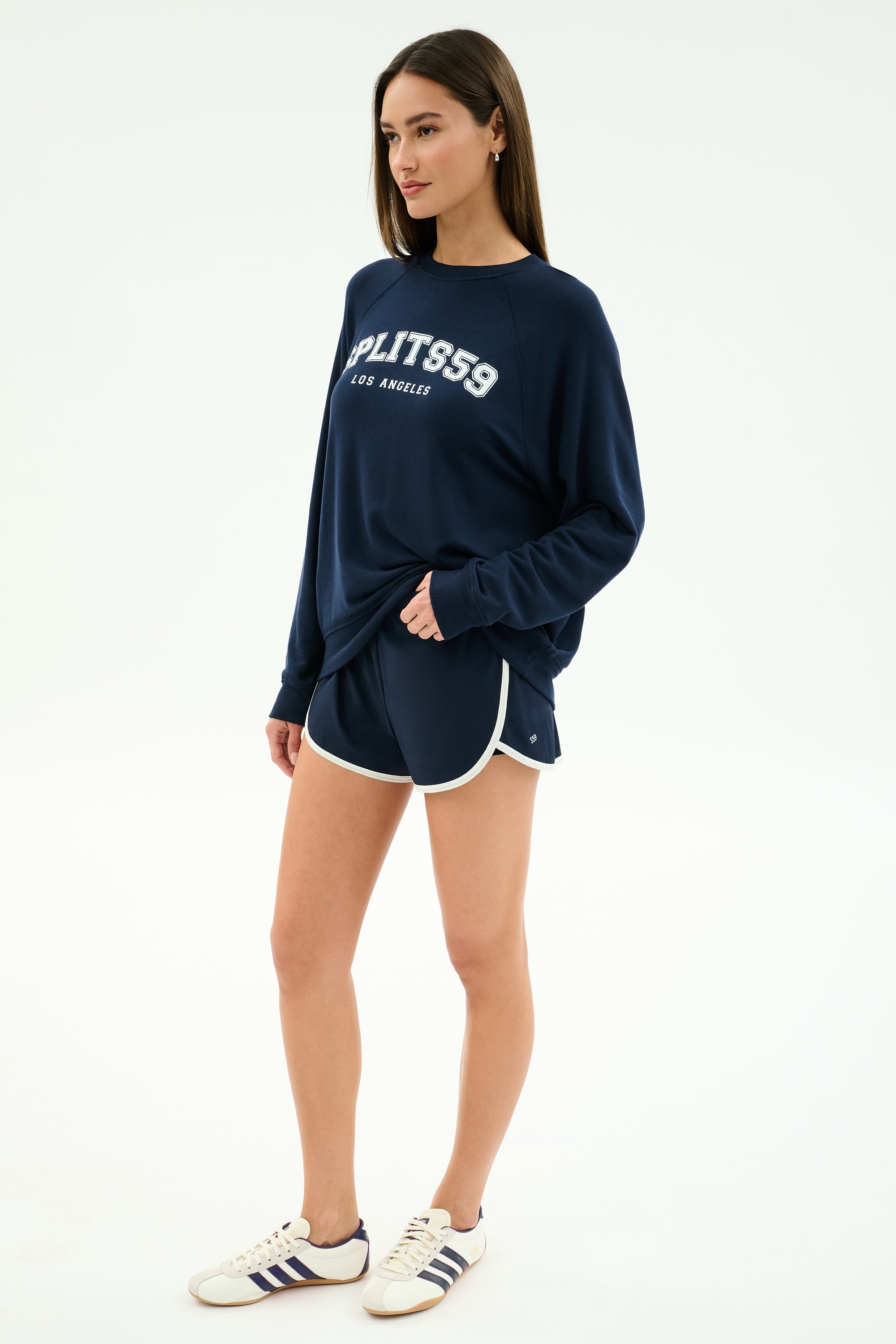 A woman stands against a plain background wearing the SPLITS59 Andie Oversized Fleece Varsity Graphic Sweatshirt in Indigo, featuring "Splits59 Los Angeles" on the front, paired with navy athletic shorts and white sneakers with navy stripes.