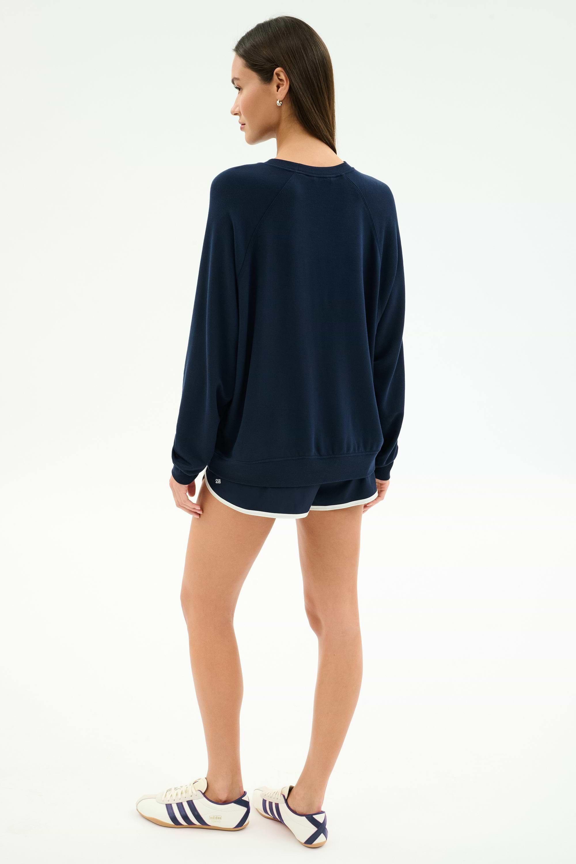 A person faces away wearing the SPLITS59 Andie Oversized Fleece Varsity Graphic Sweatshirt in indigo, navy shorts with white trim, and white Adidas sneakers with navy stripes.