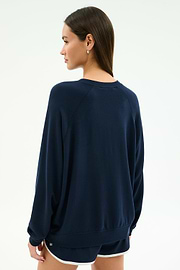 A woman stands facing away from the camera, wearing the SPLITS59 Andie Oversized Fleece Varsity Graphic Sweatshirt in Indigo and black shorts with white trim against a plain light background.