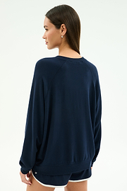 A woman stands facing away from the camera, wearing the SPLITS59 Andie Oversized Fleece Varsity Graphic Sweatshirt in Indigo and black shorts with white trim against a plain light background.