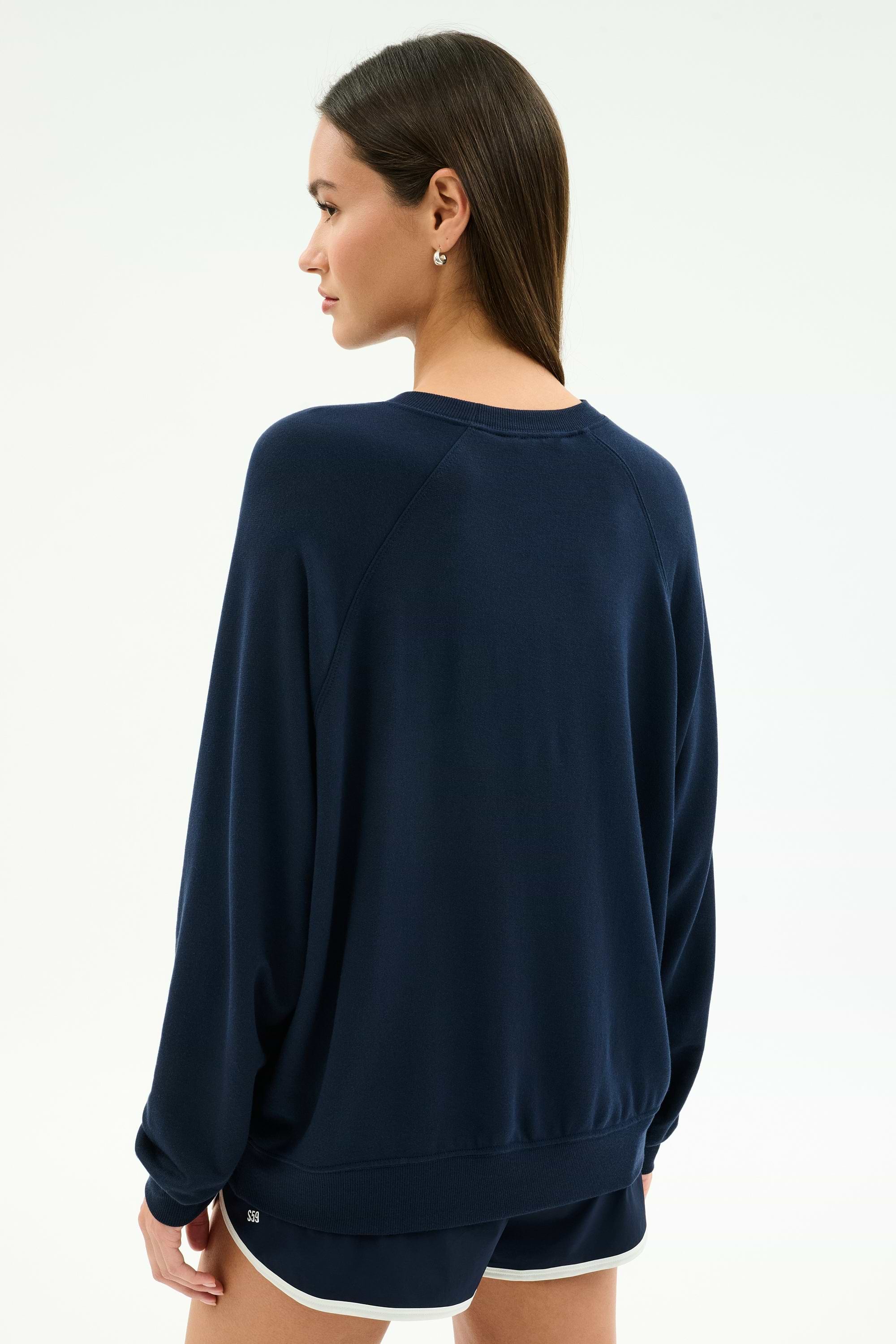 A woman stands facing away from the camera, wearing the SPLITS59 Andie Oversized Fleece Varsity Graphic Sweatshirt in Indigo and black shorts with white trim against a plain light background.