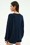 A woman stands facing away from the camera, wearing the SPLITS59 Andie Oversized Fleece Varsity Graphic Sweatshirt in Indigo and black shorts with white trim against a plain light background.