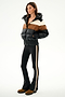 A woman wears the SPLITS59 Aspen Puffer Jacket - Black/Toffee, black flared pants with a tan side stripe, and brown shoes, posed against a plain white background.