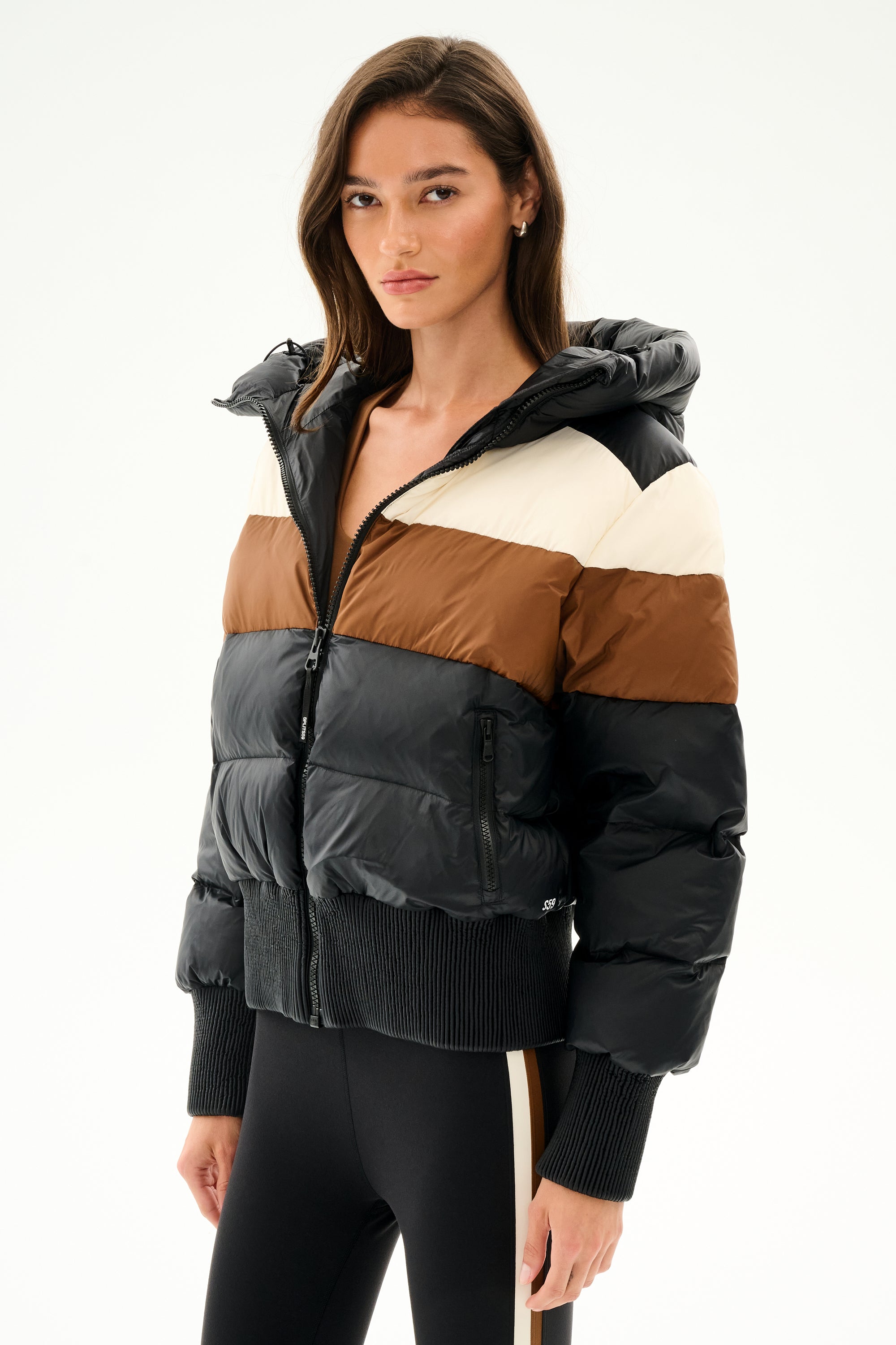 A woman wearing the Aspen Puffer Jacket - Black/Toffee by SPLITS59 and black side-striped leggings stands against a plain white background.