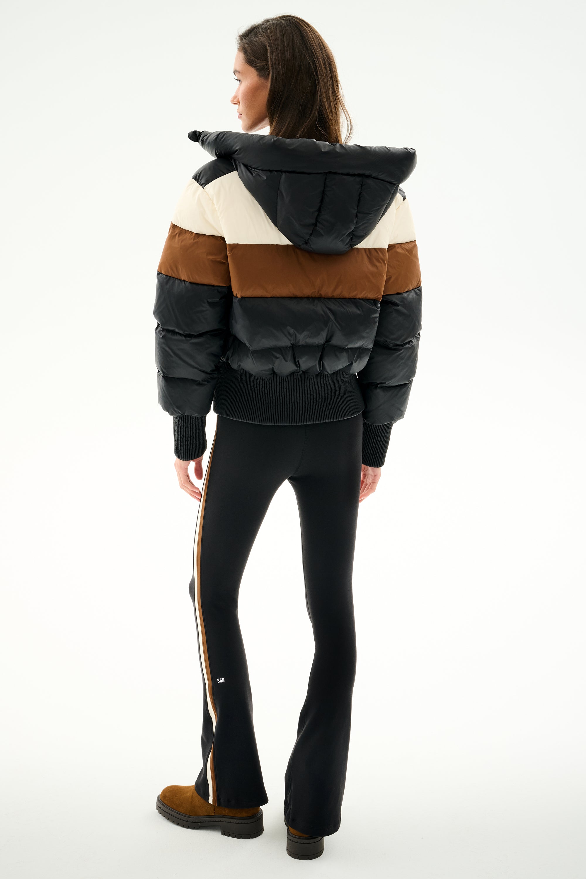 Woman faces away wearing SPLITS59 Aspen Puffer Jacket - Black/Toffee, black side-stripe pants, and brown shoes against a plain background.