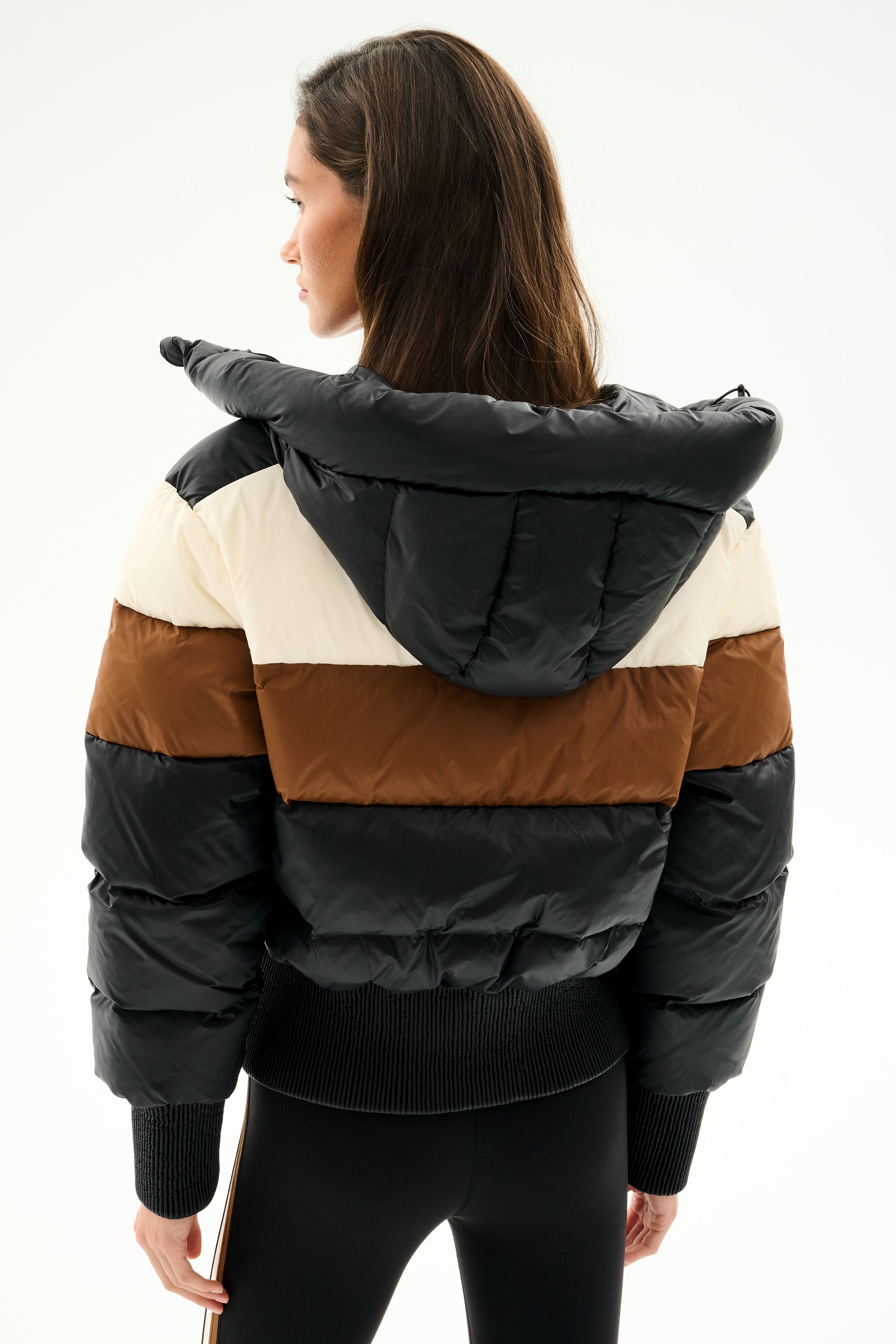 Woman facing away in the SPLITS59 Aspen Puffer Jacket - Black/Toffee, a nylon color-blocked puffer with black, brown, and cream stripes, filled with white duck down, paired with black pants.