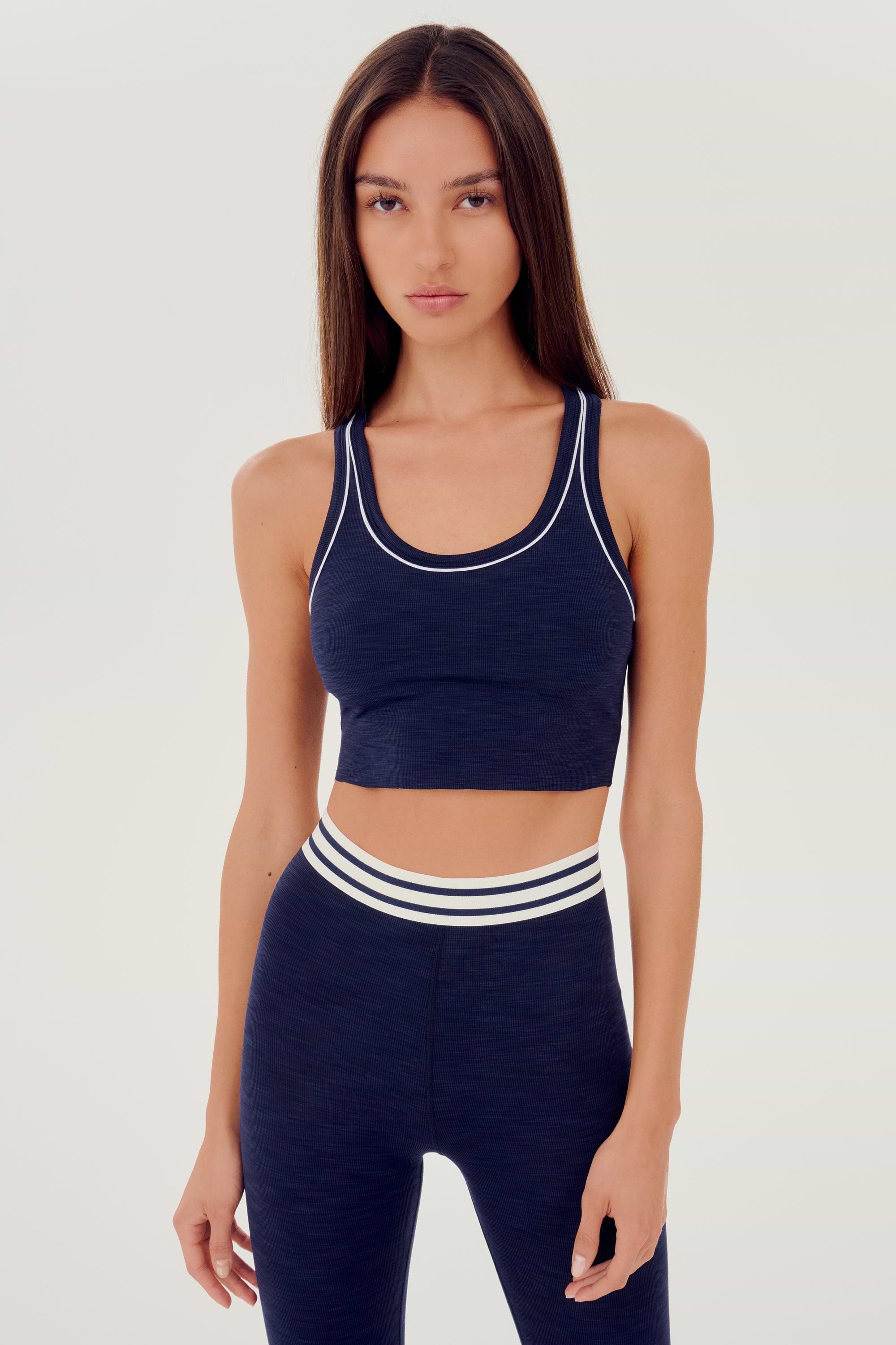 Front view of girl wearing a dark blue ribbed bra that stops at the ribs with thin white line along arms and collar with blue leggings with white and blue waistband 