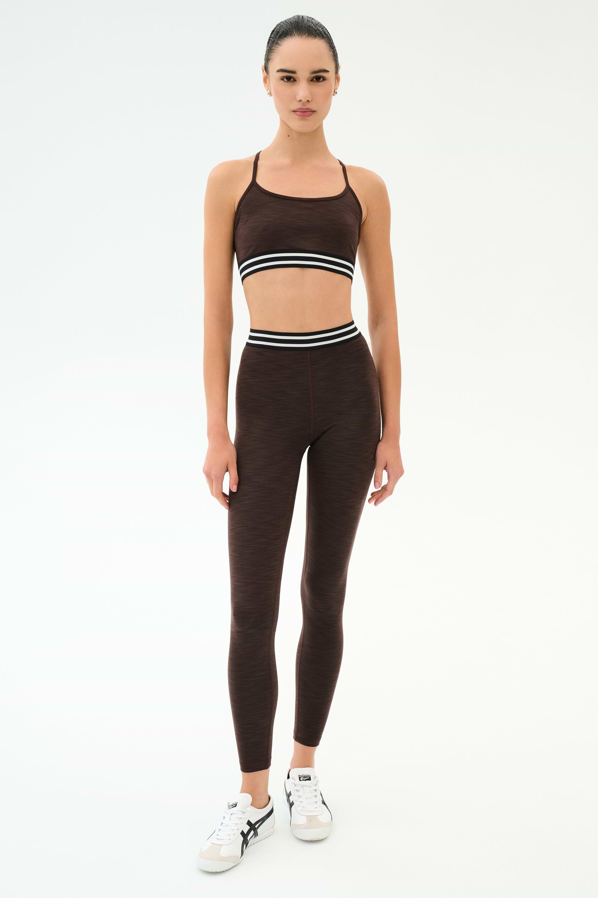 A woman wears the SPLITS59 Cali Active Rib Bra in Heather Dark Chocolate with matching leggings, featuring white and black striped waistbands, standing against a plain white background.