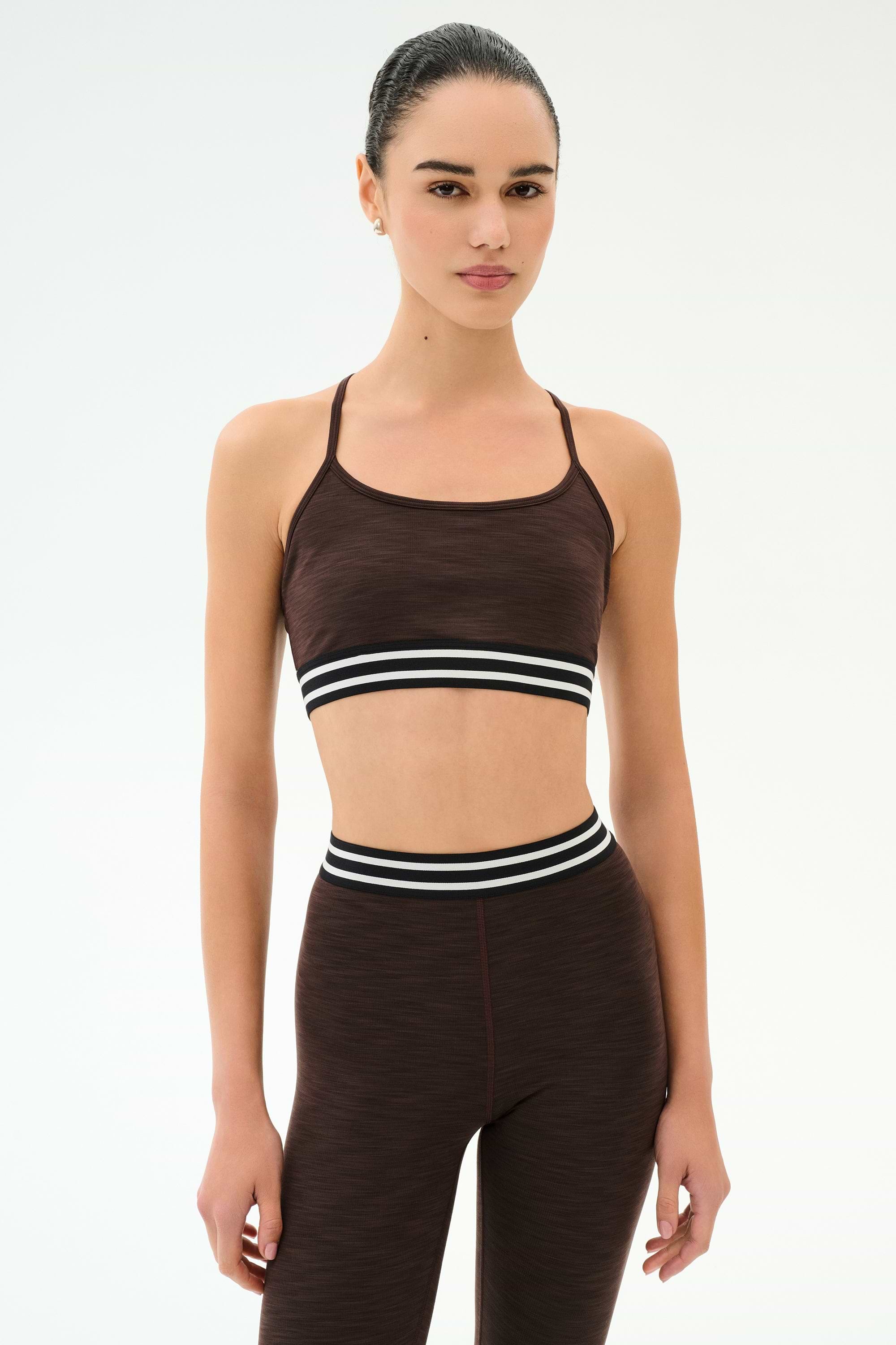 A person stands facing forward, wearing the SPLITS59 Cali Active Rib Bra in Heather Dark Chocolate and matching high-waisted leggings with black and white striped bands.